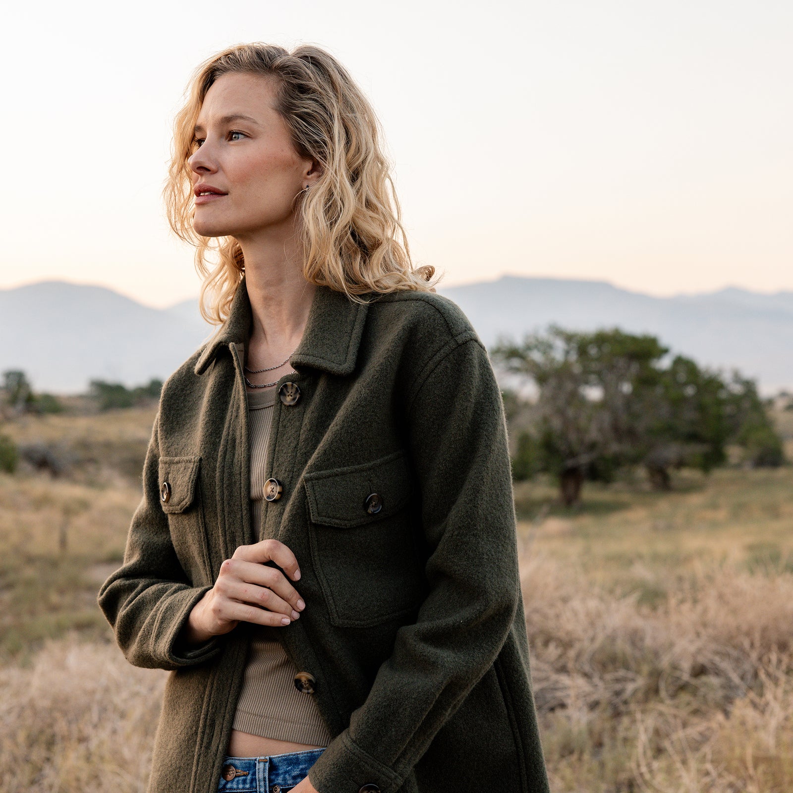 A woman with wavy blonde hair stands outdoors in a grassy landscape with distant mountains. She wears a dark green Cozy Earth Women's Boucle Shacket over a beige top and gazes to the left, her hand near the jacket's buttons. The sky is pale, and the scene has a serene, natural ambiance. 