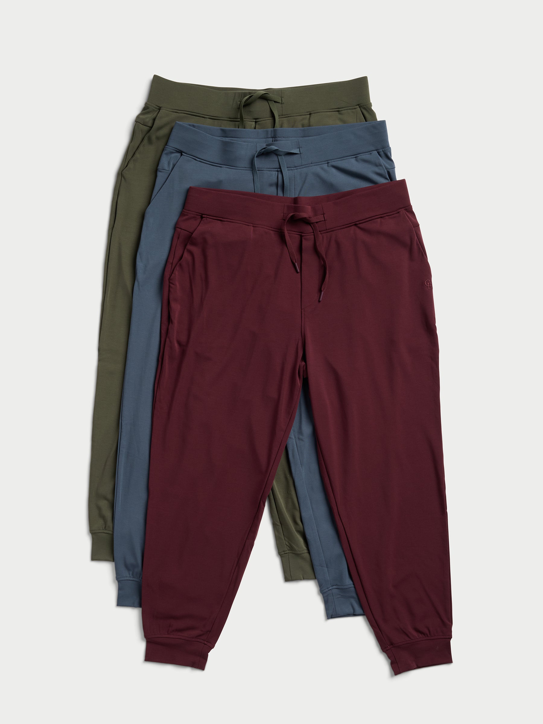 Three pairs of jogger pants are neatly arranged, overlapping each other. The colors from top to bottom are olive green, navy blue, and maroon. Each pair is designed with a drawstring waistband and cuffed ankles from Cozy Earth's Men's Ultra-Soft Bamboo Pullover Crew collection. 