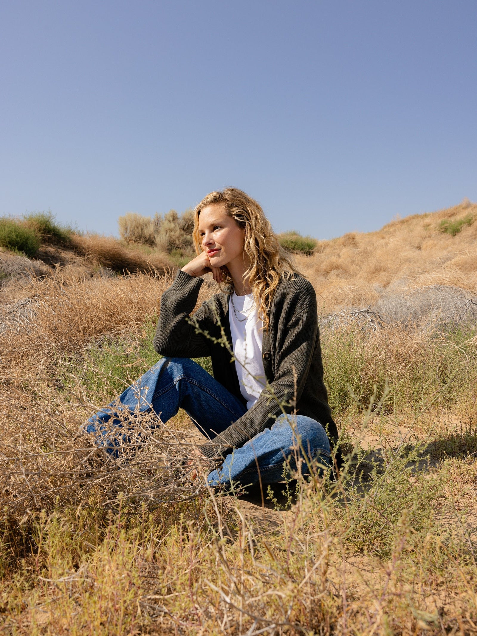 A person with curly hair, wearing a white shirt, Cozy Earth's Oversized Classic Cardigan in green, and jeans, is sitting thoughtfully on a grassy hillside under a clear blue sky. The scene is serene, with dry grass and sparse bushes in the background. 