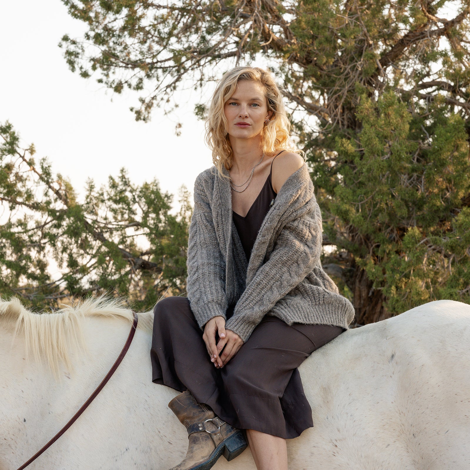 A person with blonde hair sits on a white horse, dressed in a brown dress and an Oversized Cable Knit Cardigan by Cozy Earth. They are surrounded by green trees in a natural setting, creating a calm and serene atmosphere. 