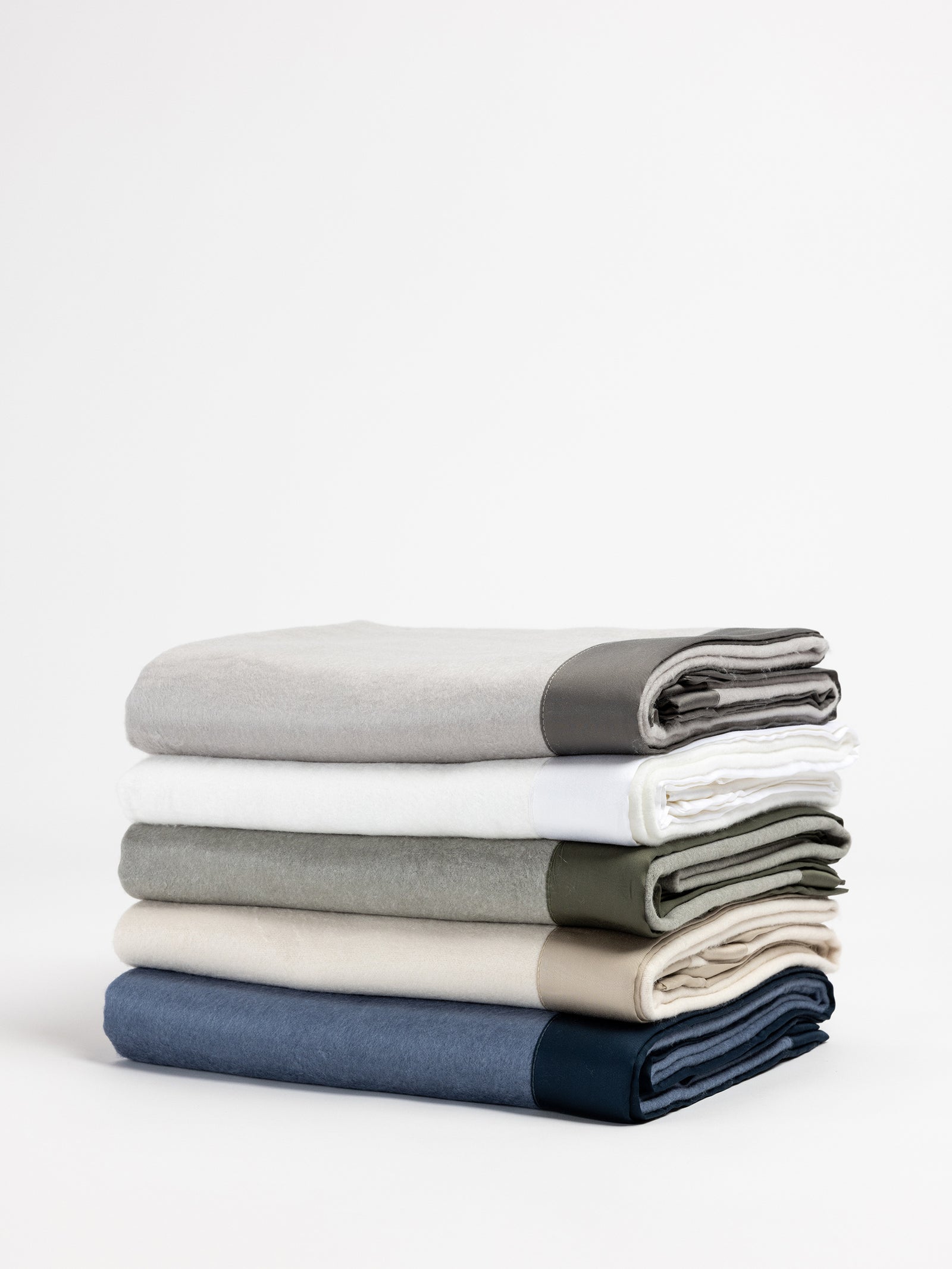 A neatly stacked pile of five folded Cozy Earth Bamboo Blankets in various colors including light gray, white, olive green, gray, and navy blue, each with a contrasting dark band at one end, set against a plain white background.