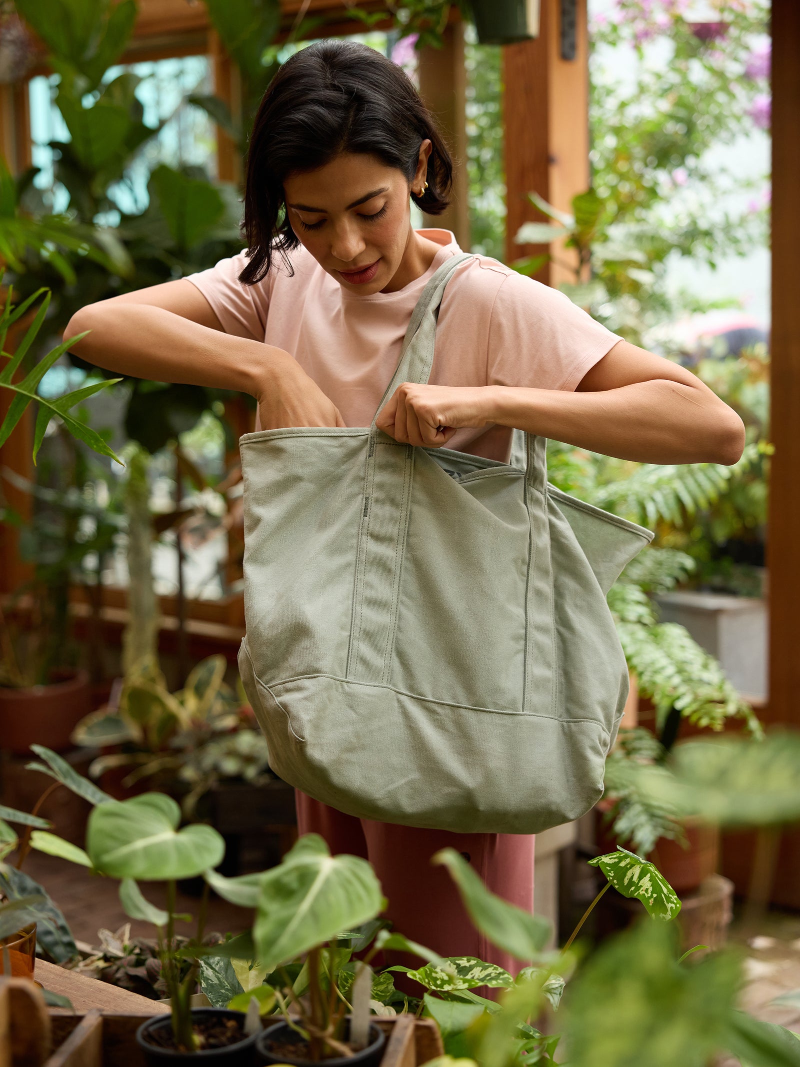 A person wearing a light pink shirt inspects the contents of a large Waxed Canvas Tote by Cozy Earth, which is light green in color. They are standing in a lush, indoor garden filled with various green plants. The ambiance is serene and filled with natural light from large windows. 