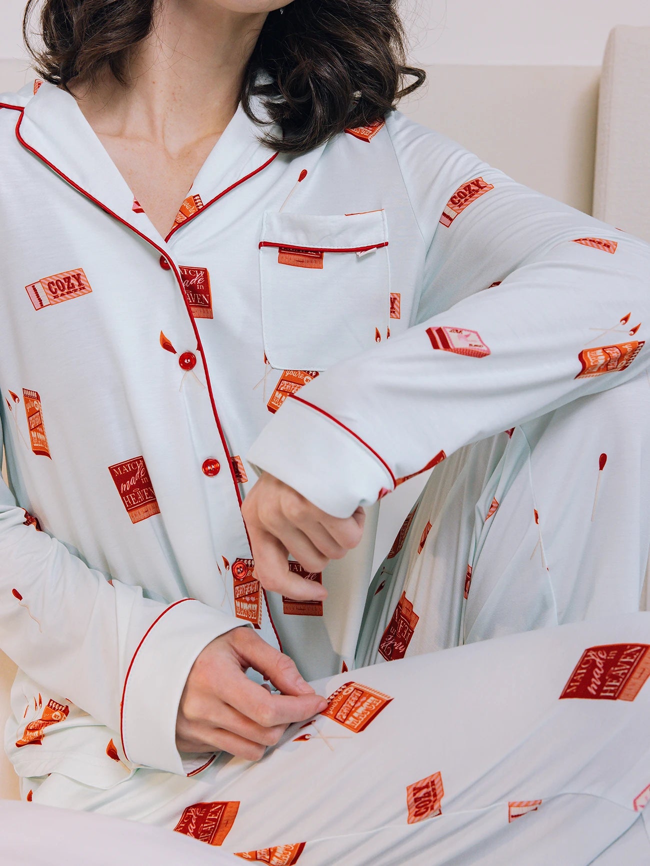 A person sits with hands on their lap in light-colored Women's Stretch-Knit Bamboo Pajama Pants by Cozy Earth, featuring red match-themed prints, buttons, and piping on the collar, sleeves, and pocket. 