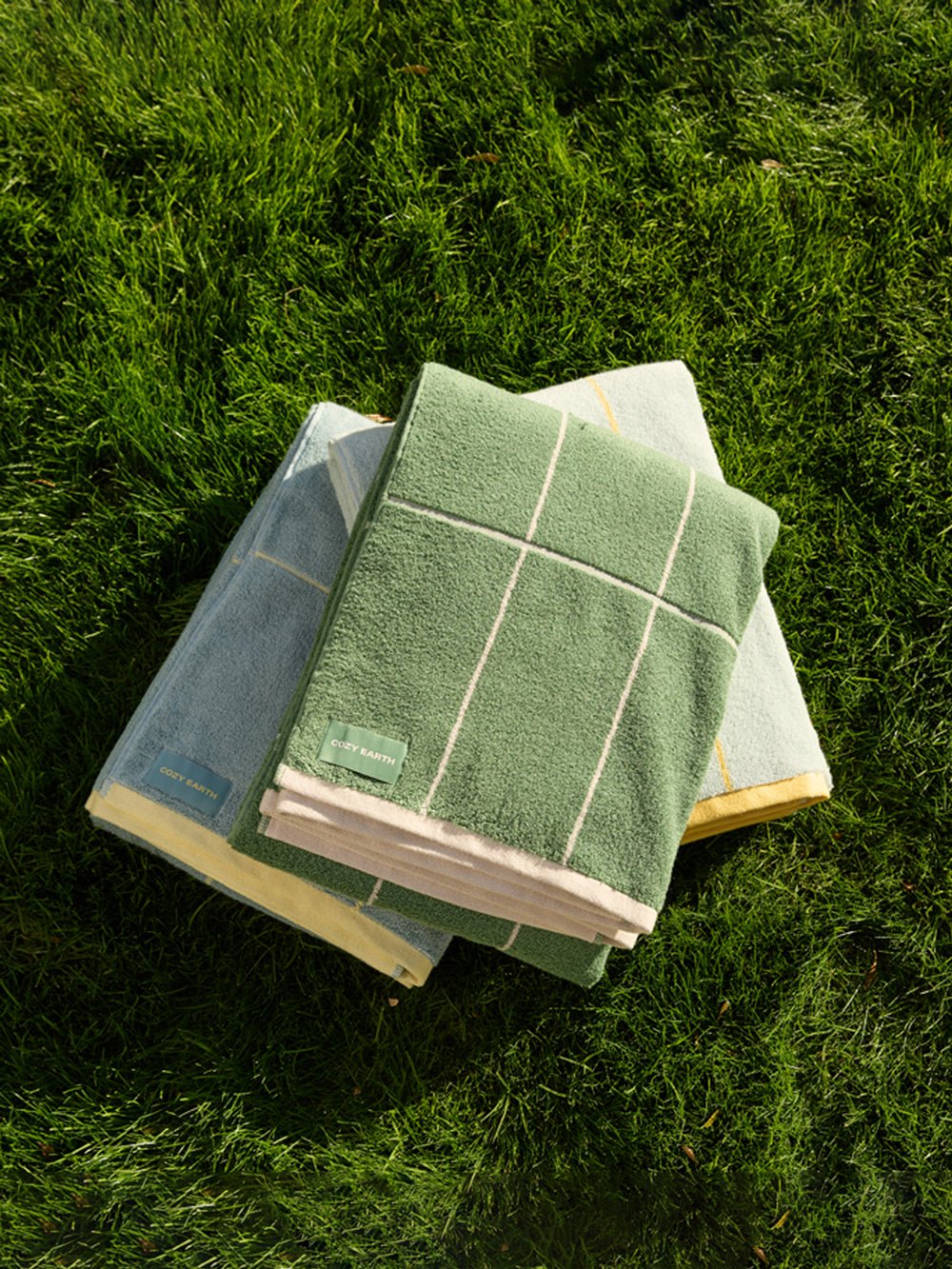 Three folded Cozy Earth Windowpane Resort Towels, in green, blue, and light yellow with small green labels attached, rest on lush grass. Sunlight creates shadows that accentuate the texture of both the towels and the grass. 