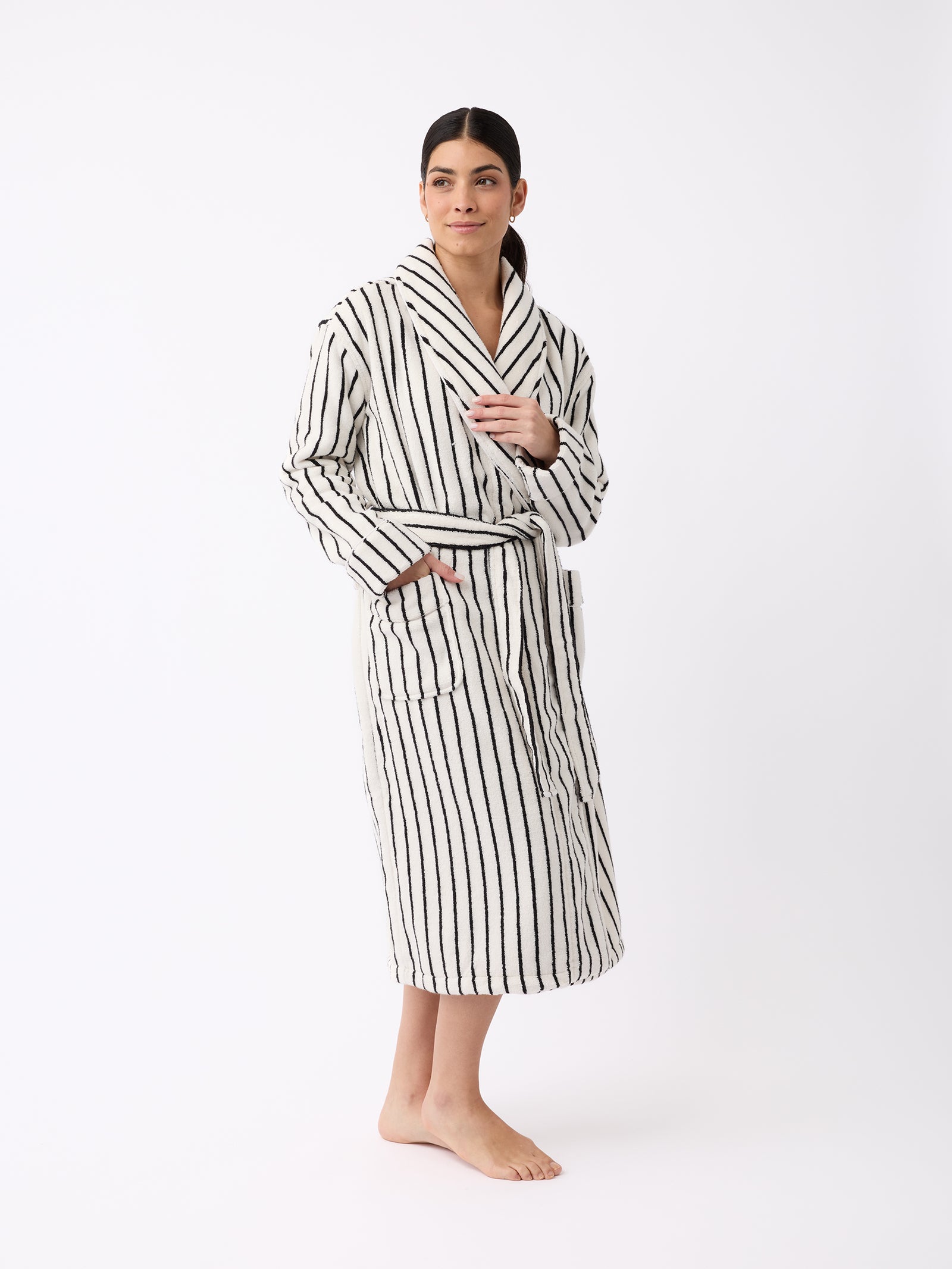 A person models the Luxe Bath Robe by Cozy Earth, featuring black vertical stripes on a white fabric. They stand barefoot against a white background, gazing to the side while one hand holds the robe's collar and the other rests on its belt tie. 