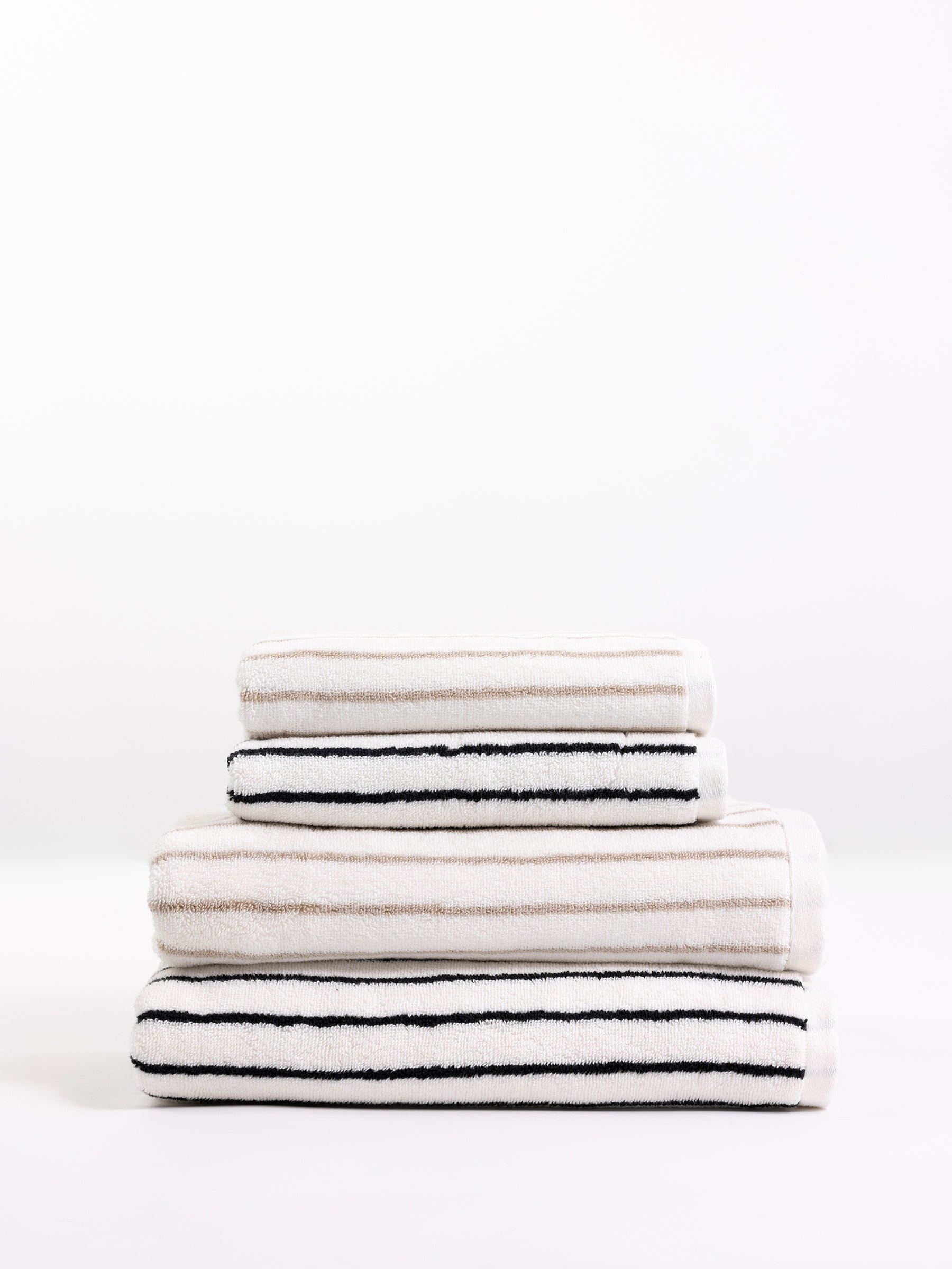 A stack of Cozy Earth's Luxe Bath Towels, neatly folded in varying sizes and arranged in a pyramid shape. The towels feature a white background with horizontal black and beige stripes, providing a modern and minimalistic look. The backdrop is plain and white. 