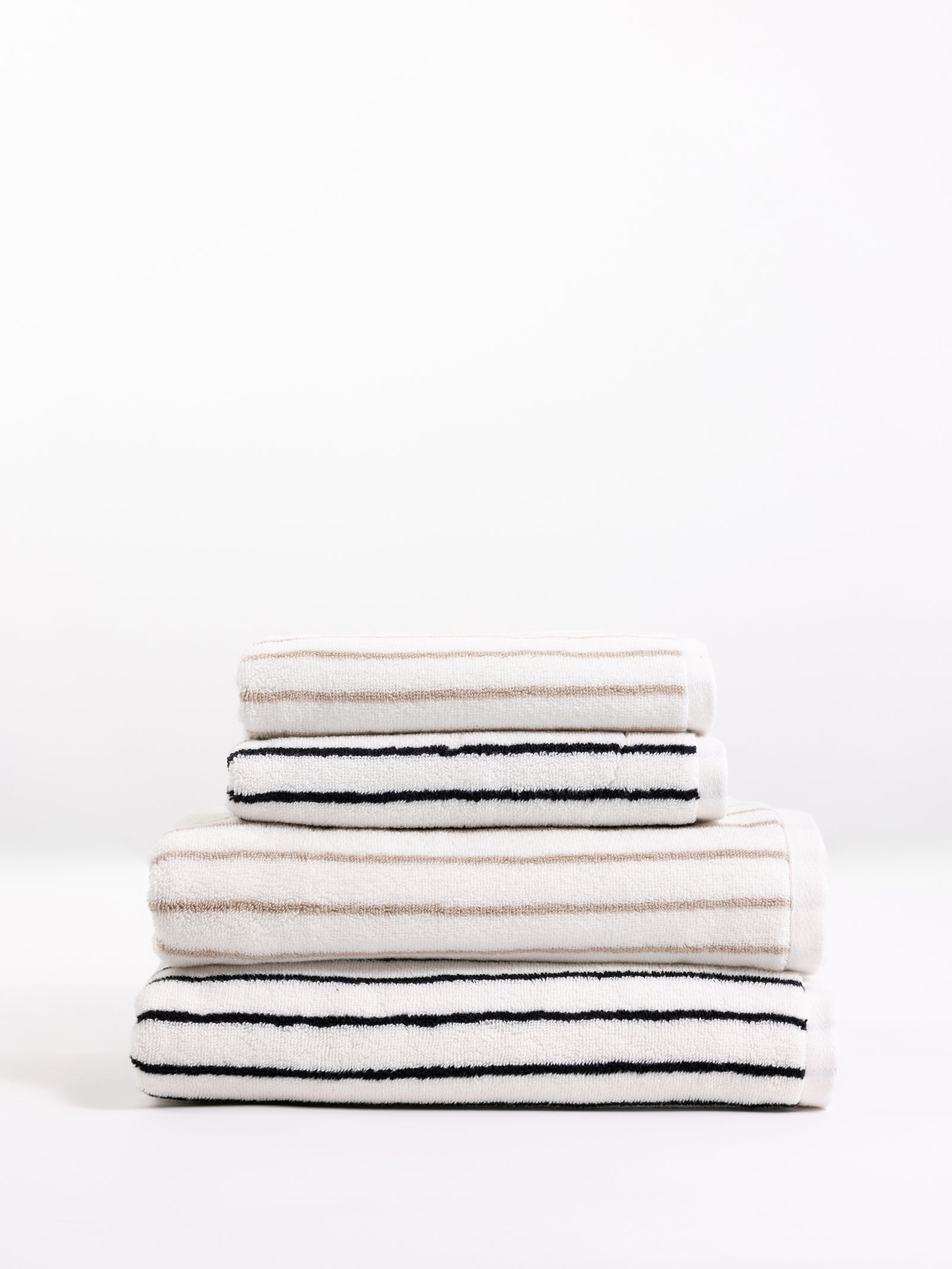 A stack of Cozy Earth's Luxe Bath Towels, neatly folded in varying sizes and arranged in a pyramid shape. The towels feature a white background with horizontal black and beige stripes, providing a modern and minimalistic look. The backdrop is plain and white. 