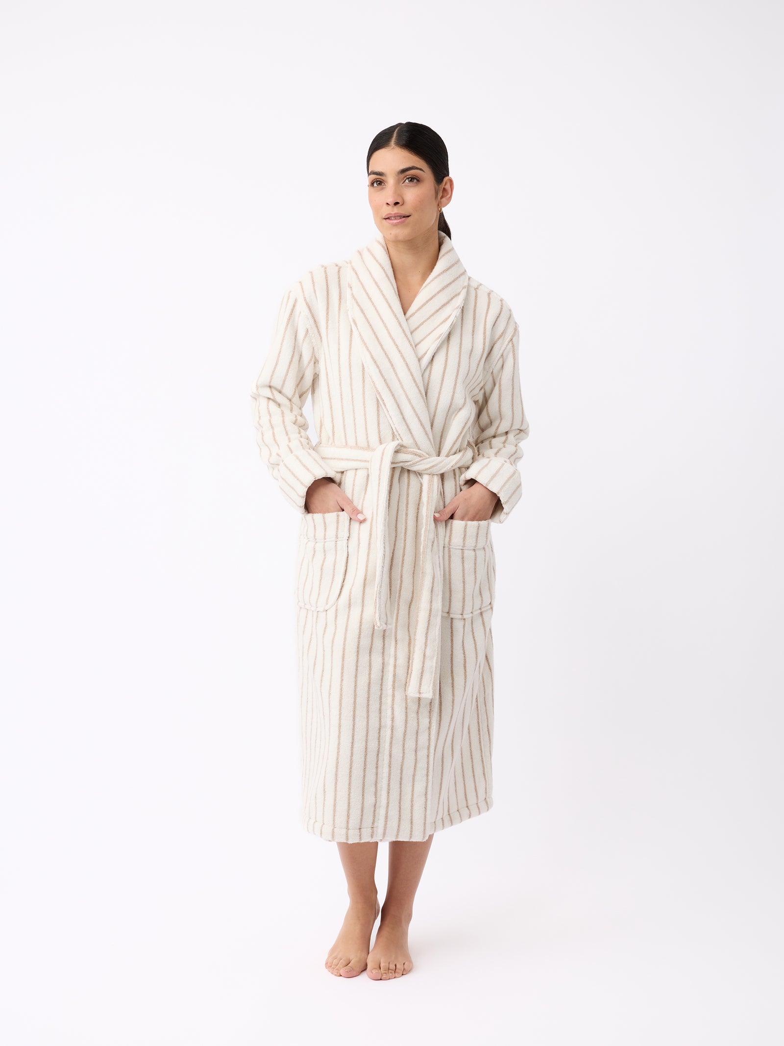 A person stands barefoot wearing the Luxe Bath Robe by Cozy Earth, featuring a cream color with vertical tan stripes. The robe is belted at the waist and includes two large pockets. The background is plain white. 