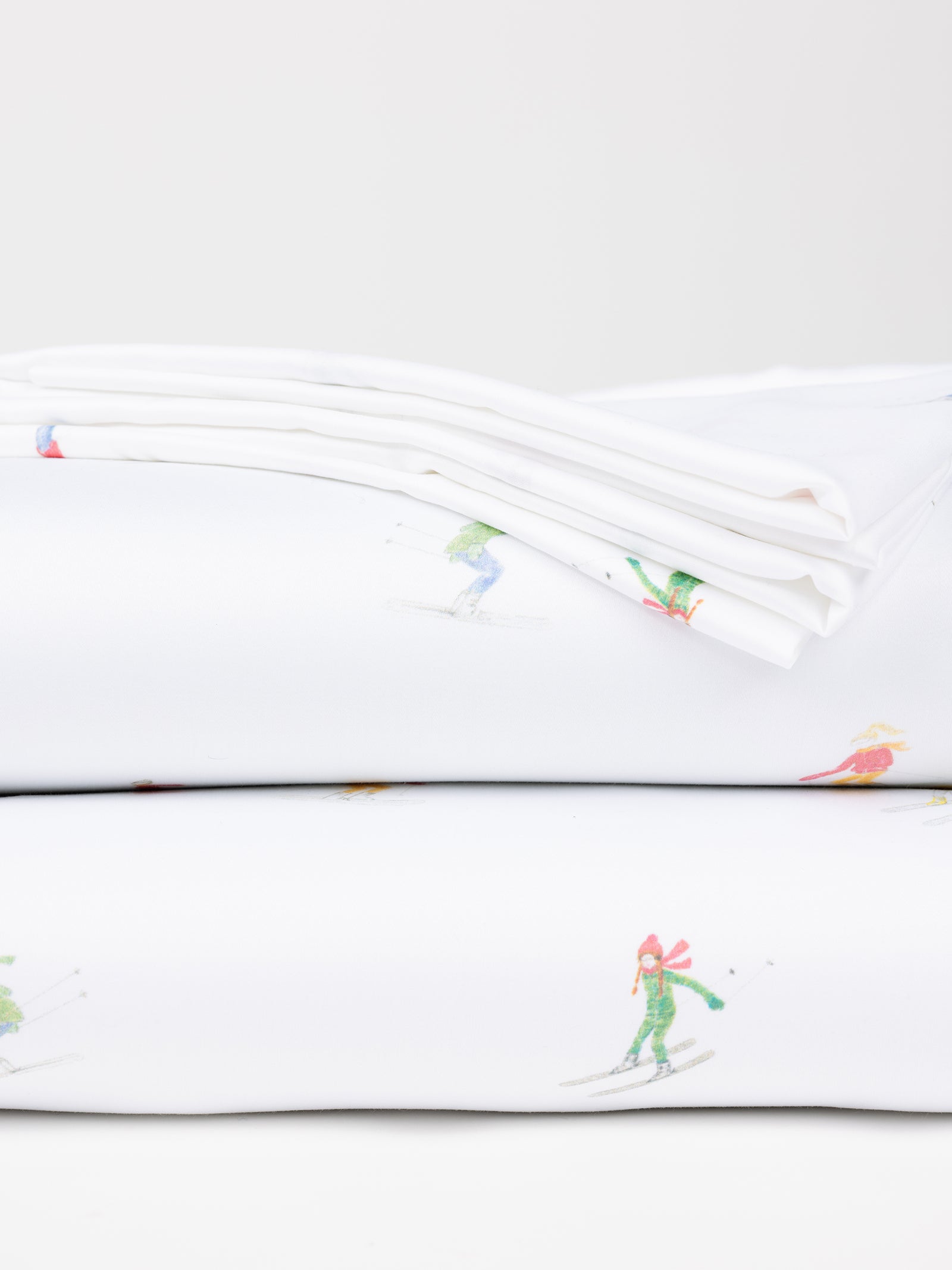 Folded Bamboo Sheet Set in white, from Cozy Earth, featuring a subtle pattern of small, colorful skiing figures, stacked on top of each other against a plain white background. 