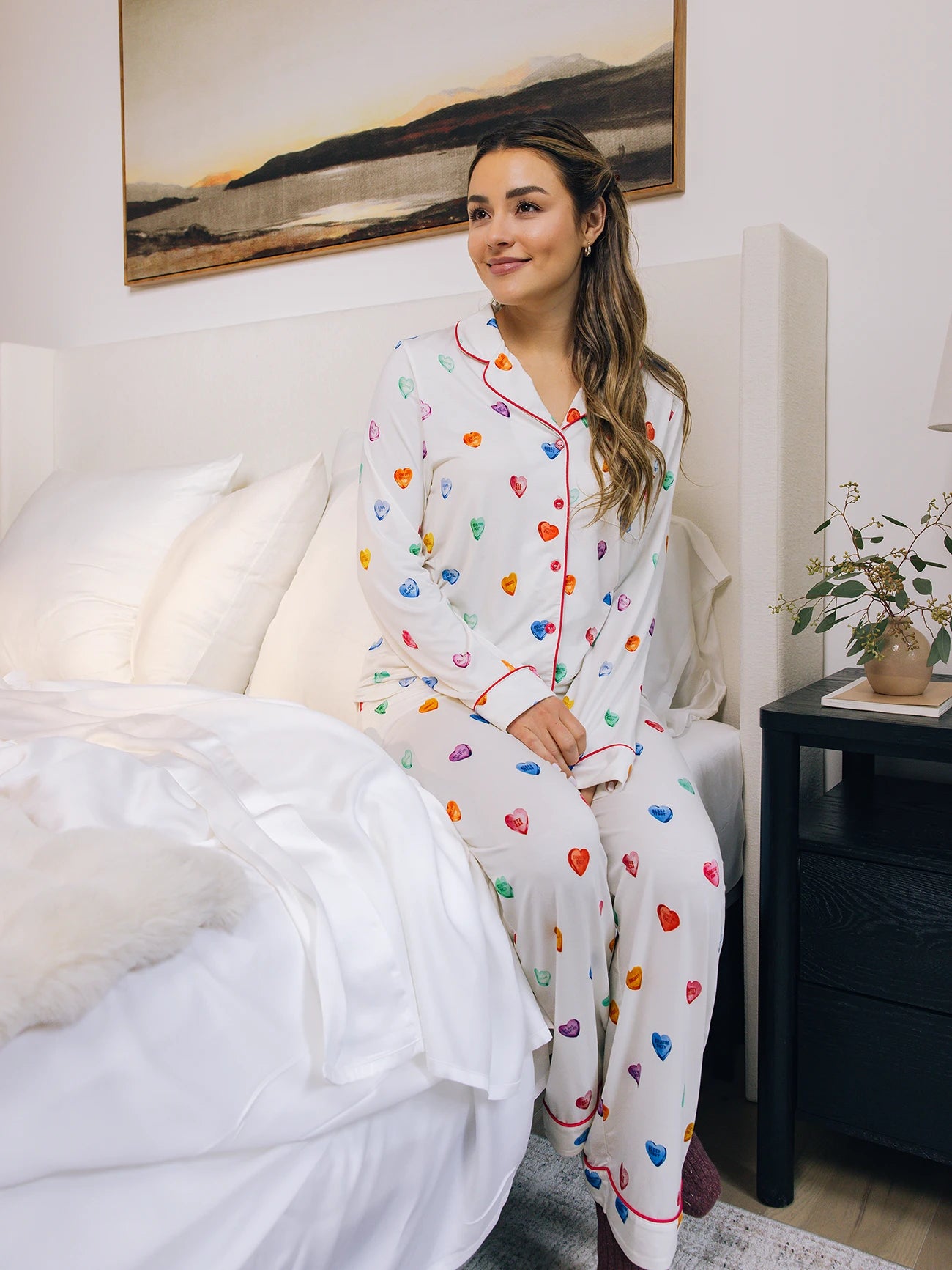 A woman sits on a bed wearing Cozy Earth Women's Stretch-Knit Bamboo Pajama Pant with colorful heart patterns, smiling gently. Nearby, there's a bedside table with a plant and lamp, beneath a mountain landscape picture on the wall. 