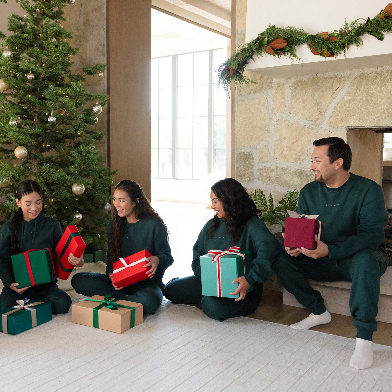 A family of four, dressed in matching green Men's CityScape Crewnecks by Cozy Earth, sits on the floor near a beautifully decorated Christmas tree and stone fireplace. They are smiling and holding wrapped gifts in various colors, while garland elegantly decorates the mantel. 