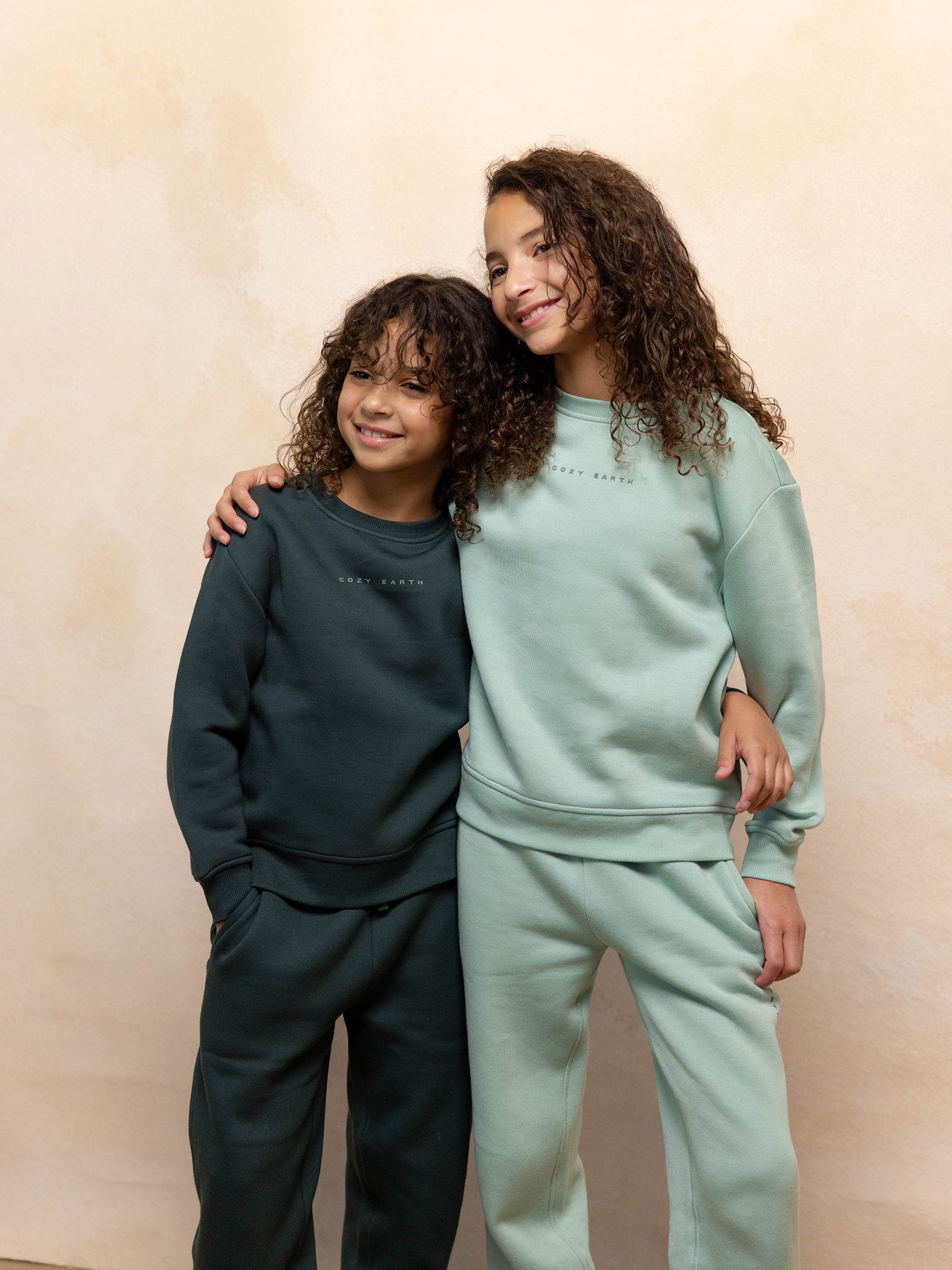 Two children with curly hair, both wearing matching outfits from Cozy Earth's Kid's CityScape Crewneck collection, smile and stand side by side with their arms around each other. One child is dressed in a dark green tracksuit, while the other wears a light green tracksuit against a neutral-toned background. 