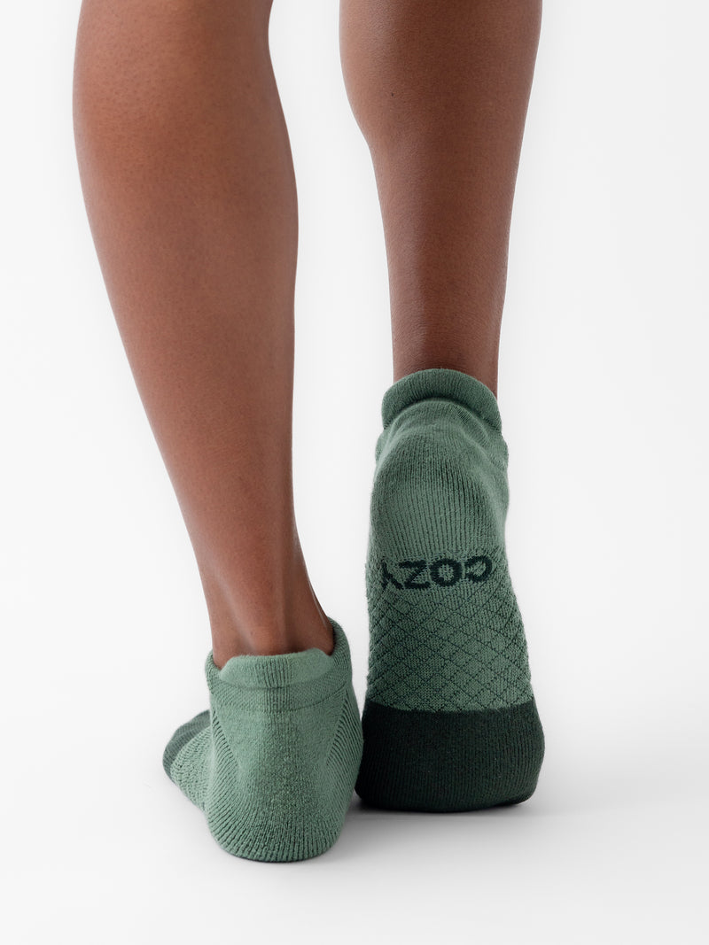 A person wearing green socks from the Cozy Earth Essential Ankle Sock 2-Pack collection, featuring a black pattern and a black strip on the heel. The word "cozy" is visible on one sock as they stand with their feet close together on a white surface.