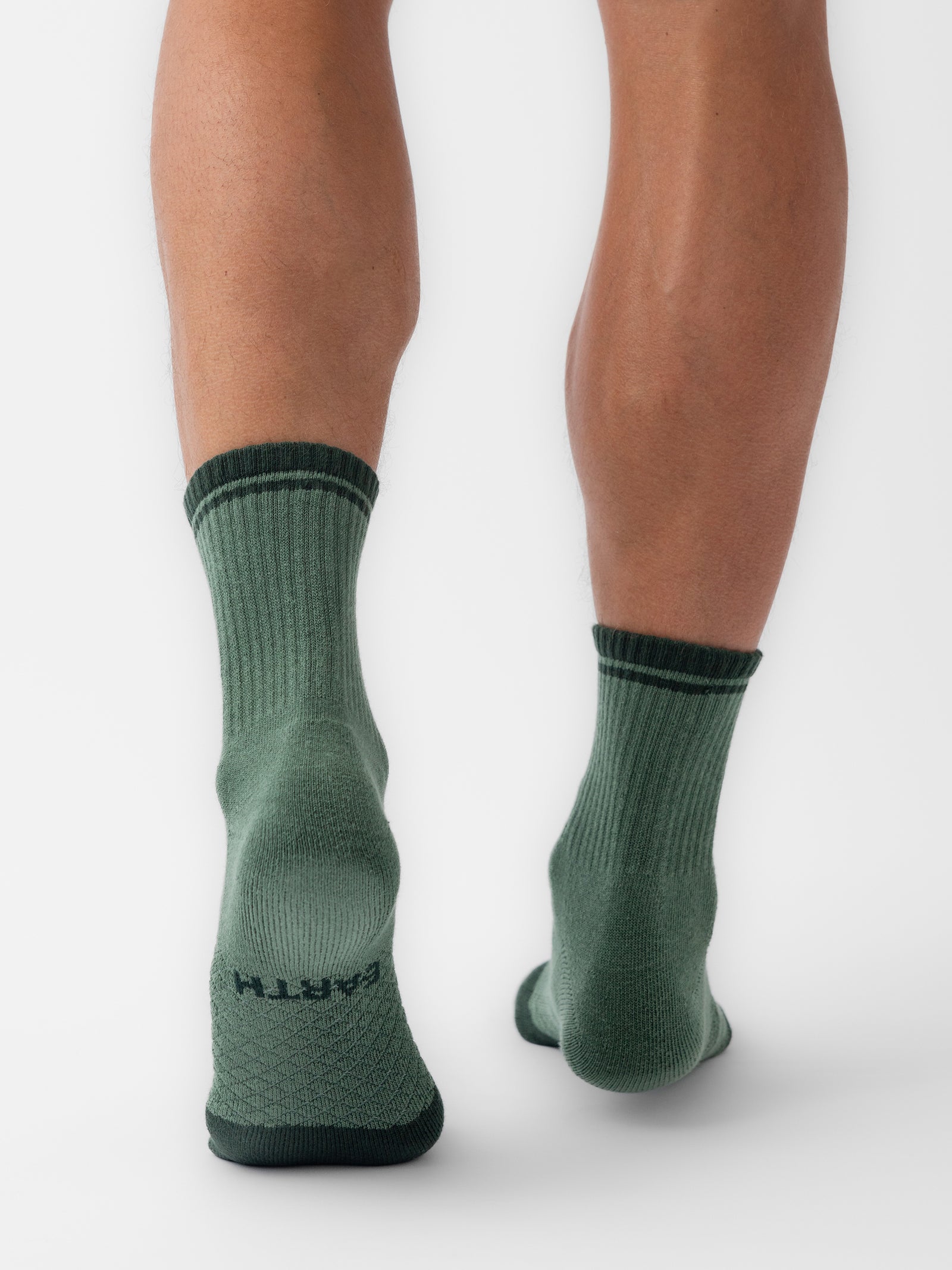 The lower legs of a person are shown, adorned in Cozy Earth's Essential Quarter Socks. The green socks feature dark green stripes at the top and dark green trim at the toes and heels, with the word "EARTH" visible on the sole of the right sock. The background is plain white. 