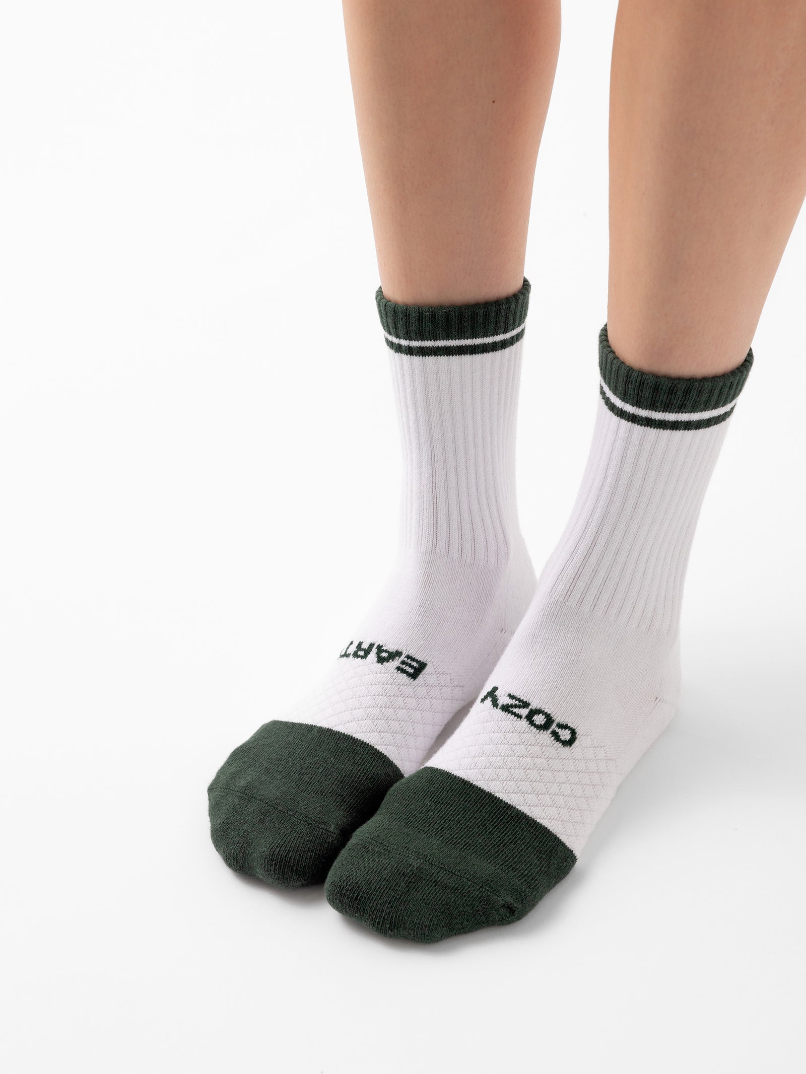 A person wearing white socks with green accents on the toes, heels, and cuffs from the Essential Calf Sock 4-Pack by Cozy Earth. The socks feature the words "COZY EARS" printed in black text near the toes. The person is standing on a white background. 