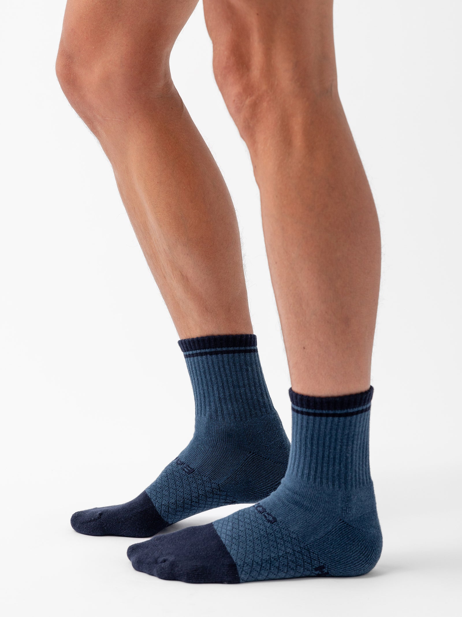 Side view of a person wearing Cozy Earth's Essential Quarter Sock from the 4-Pack in blue and navy. The ankle socks feature a ribbed cuff, darker heel and toe sections, and a patterned mid-foot area. The person is standing with one leg slightly forward, displaying the socks' fit and design. 