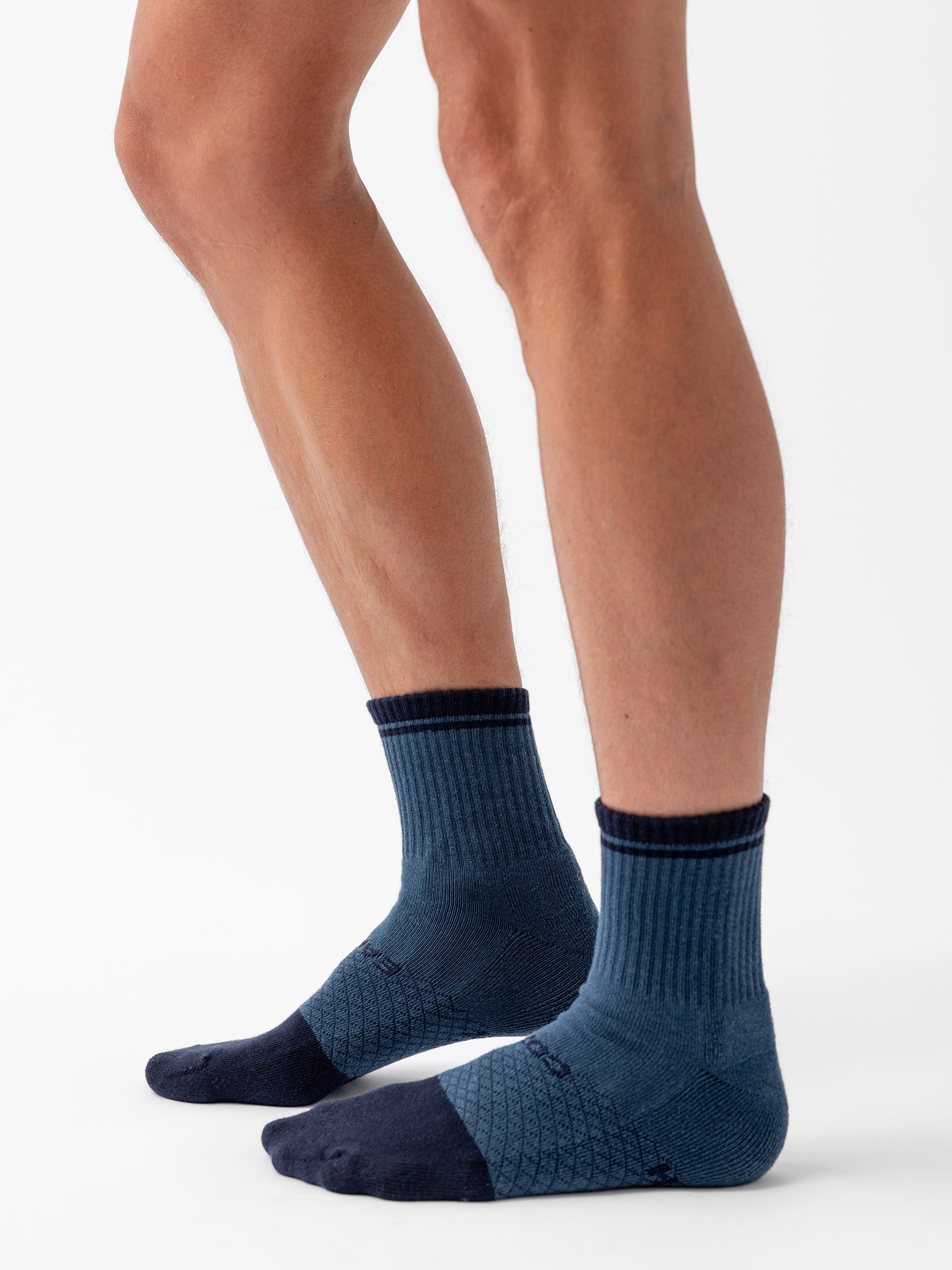 Side view of a person wearing Cozy Earth's Essential Quarter Sock from the 4-Pack in blue and navy. The ankle socks feature a ribbed cuff, darker heel and toe sections, and a patterned mid-foot area. The person is standing with one leg slightly forward, displaying the socks' fit and design. |Color:Spruce/Eclipse