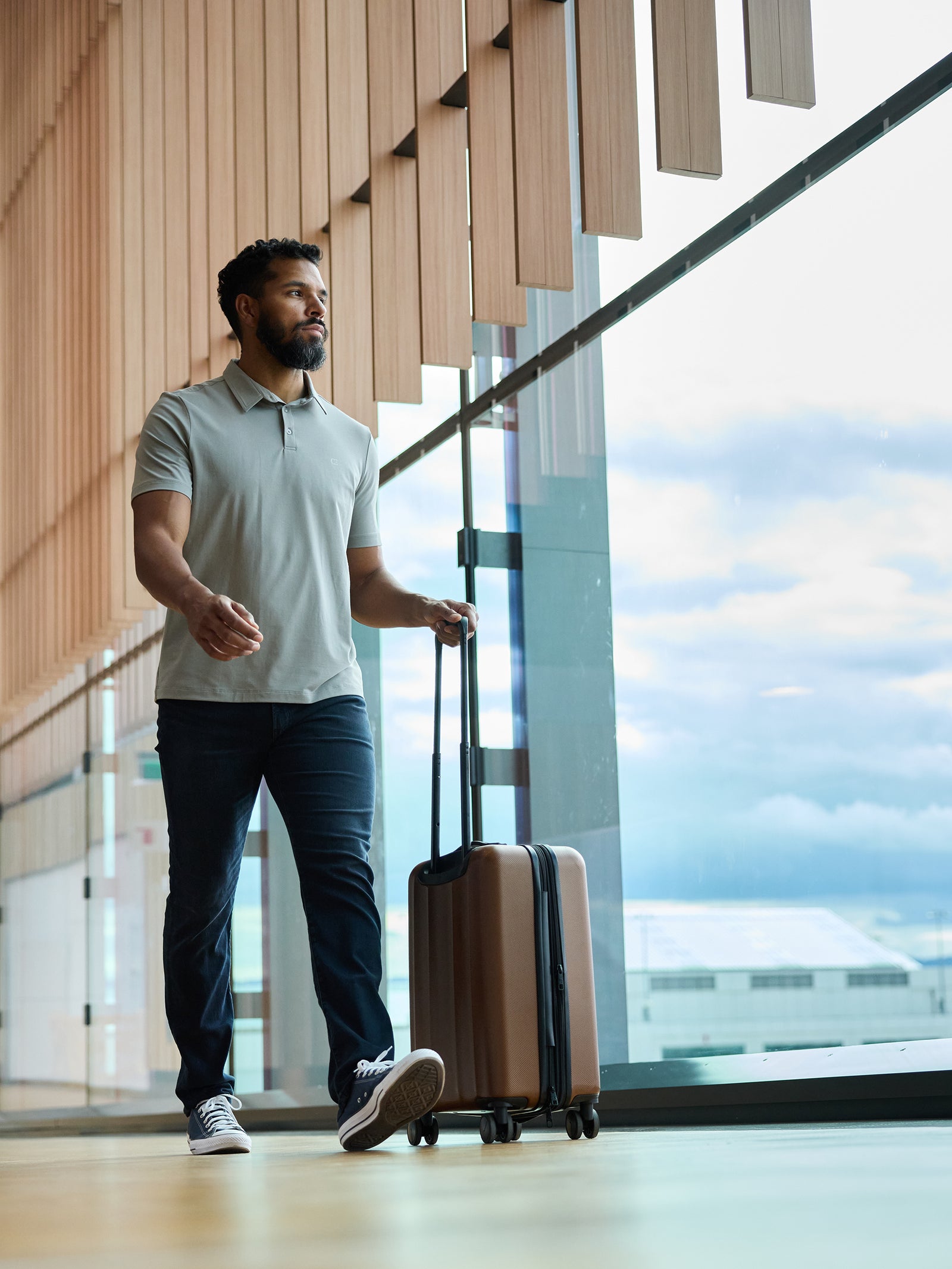 A man with a beard strolls through an airport terminal, pulling a bronze suitcase. He is wearing the Men's Everyday Polo by Cozy Earth in gray, paired with dark pants and sneakers. Large windows reveal a cloudy sky outside. 