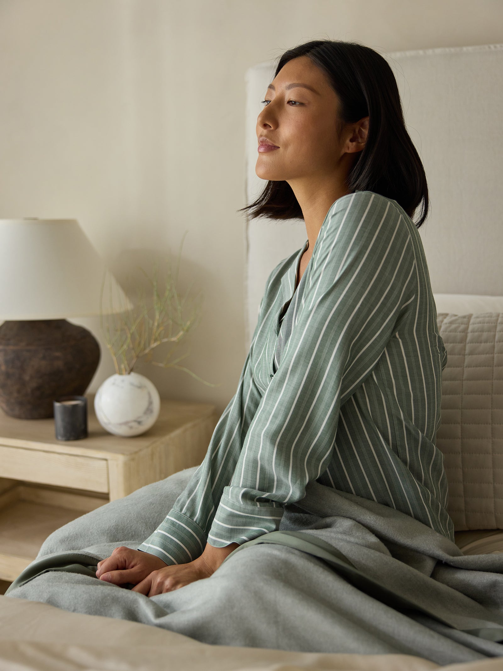 A woman with short dark hair sits on a bed, gazing thoughtfully towards a window. She wears the Cozy Earth Women's Soft Woven Long Sleeve Pajama Top in green and white stripes. A lamp and two decorative vases are on a wooden nightstand nearby. The room is decorated in a calm and neutral color palette. 