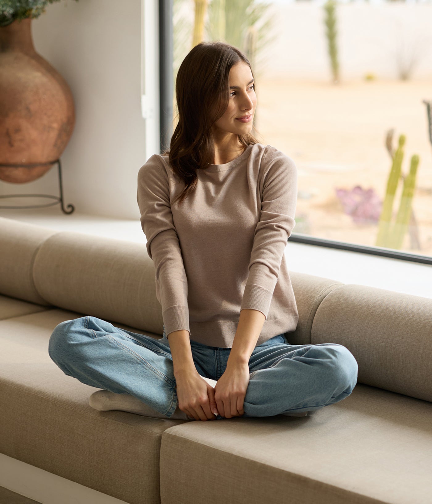 A woman with long hair sits cross-legged on a beige sofa, wearing Cozy Earth's Women's AirKnit Crewneck Sweater and blue jeans. With a calm expression, they gaze out the window. Cacti and a large clay pot are in the background. 