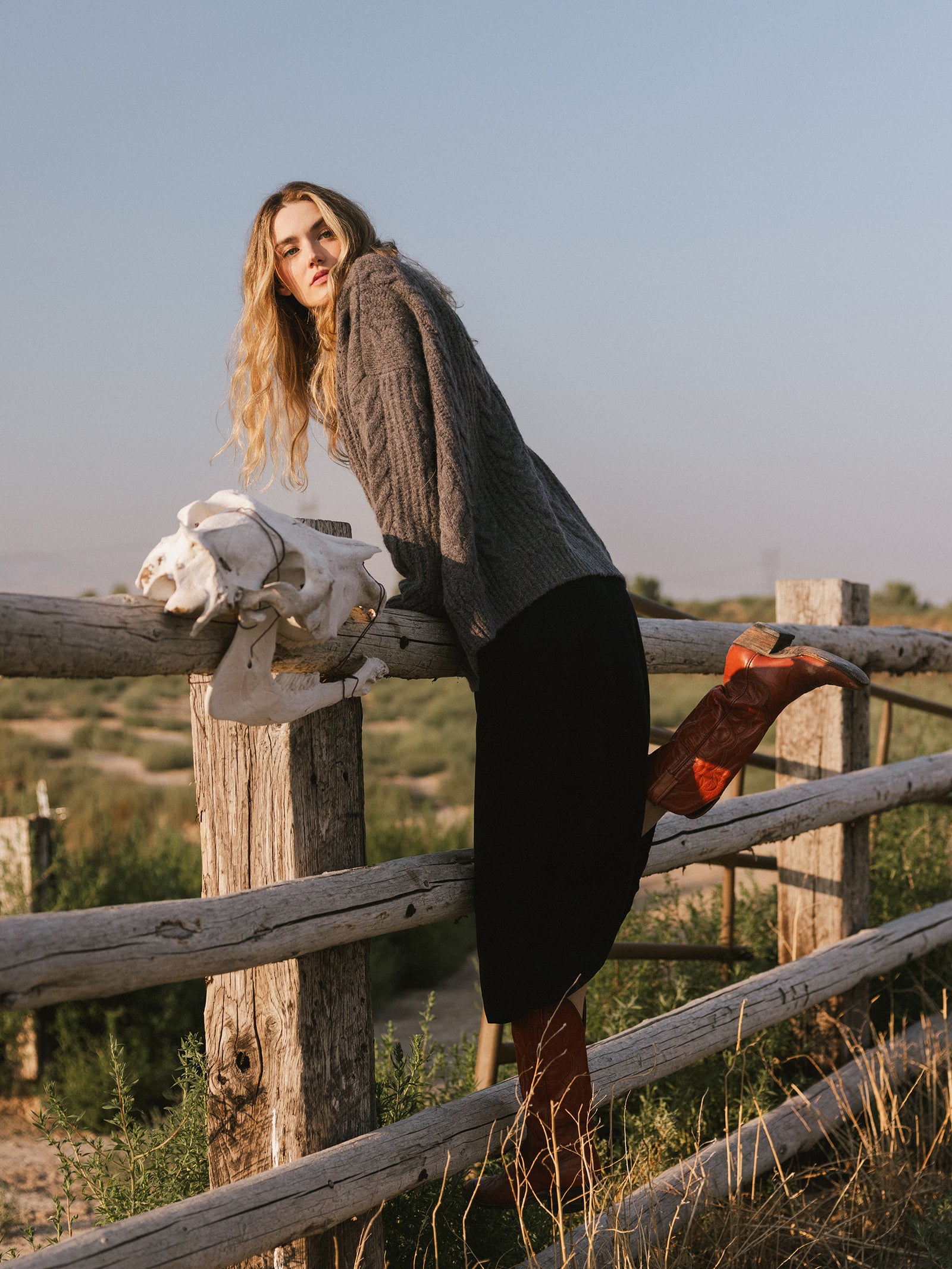 A person with long hair, dressed in an Oversized Cable Knit Cardigan by Cozy Earth, a black skirt, and red boots, leans on a wooden fence while gazing into the distance. A white skull is positioned next to them in this rural setting under a clear blue sky. 