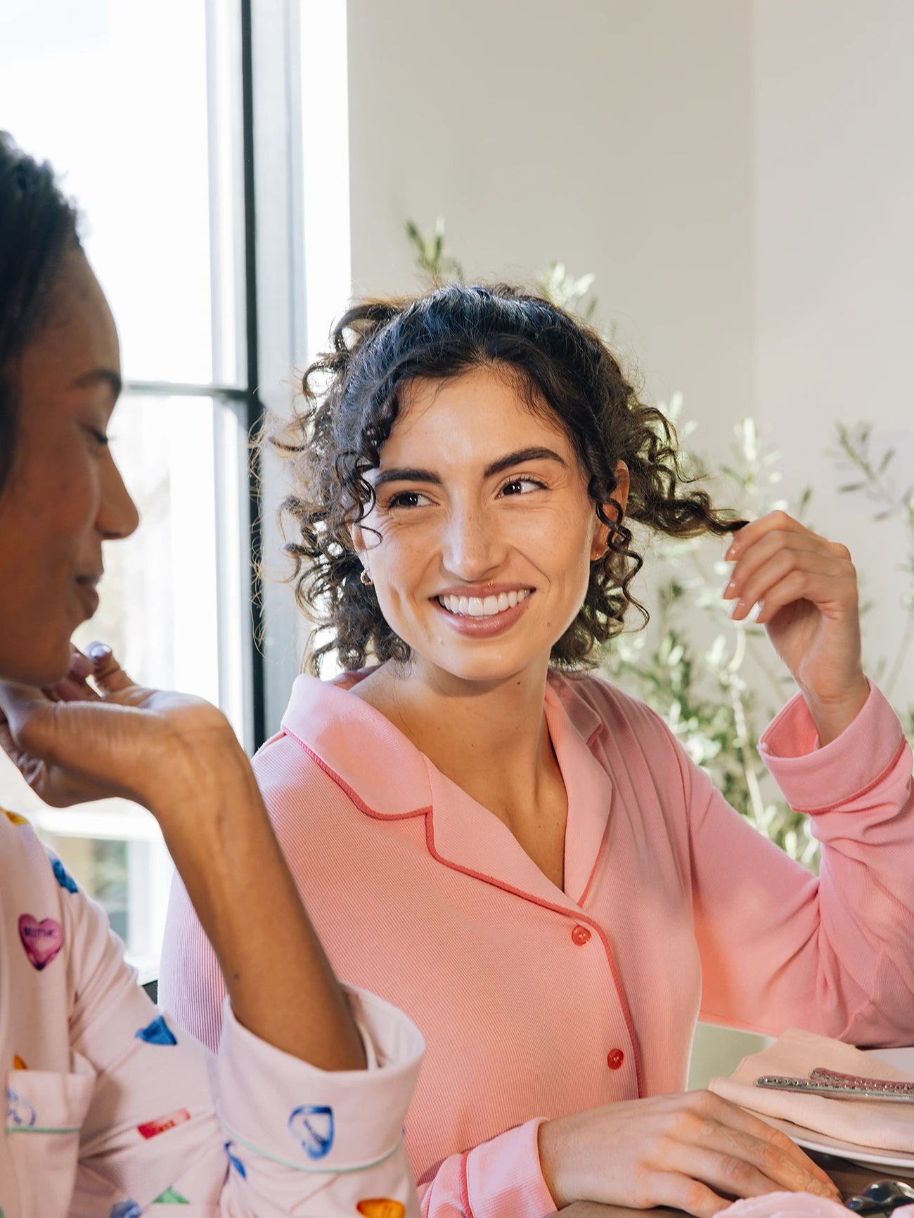 Two woman are at a table. One, in a pink Cozy Earth Women's Bamboo Rib Knit Pajama Top, is smiling while holding her hair. The other, partially visible, wears a colorful shirt. They converse as sunlight streams through the window behind them. 