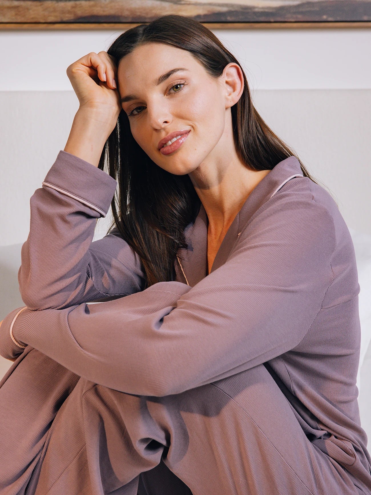 A woman with long dark hair, wearing Cozy Earth's Bamboo Rib Knit Classic Long Sleeve Pajama Top in Twilight, sits on a bed leaning her head on her hand and smiling softly. The background is a white wall with part of a picture frame visible. 