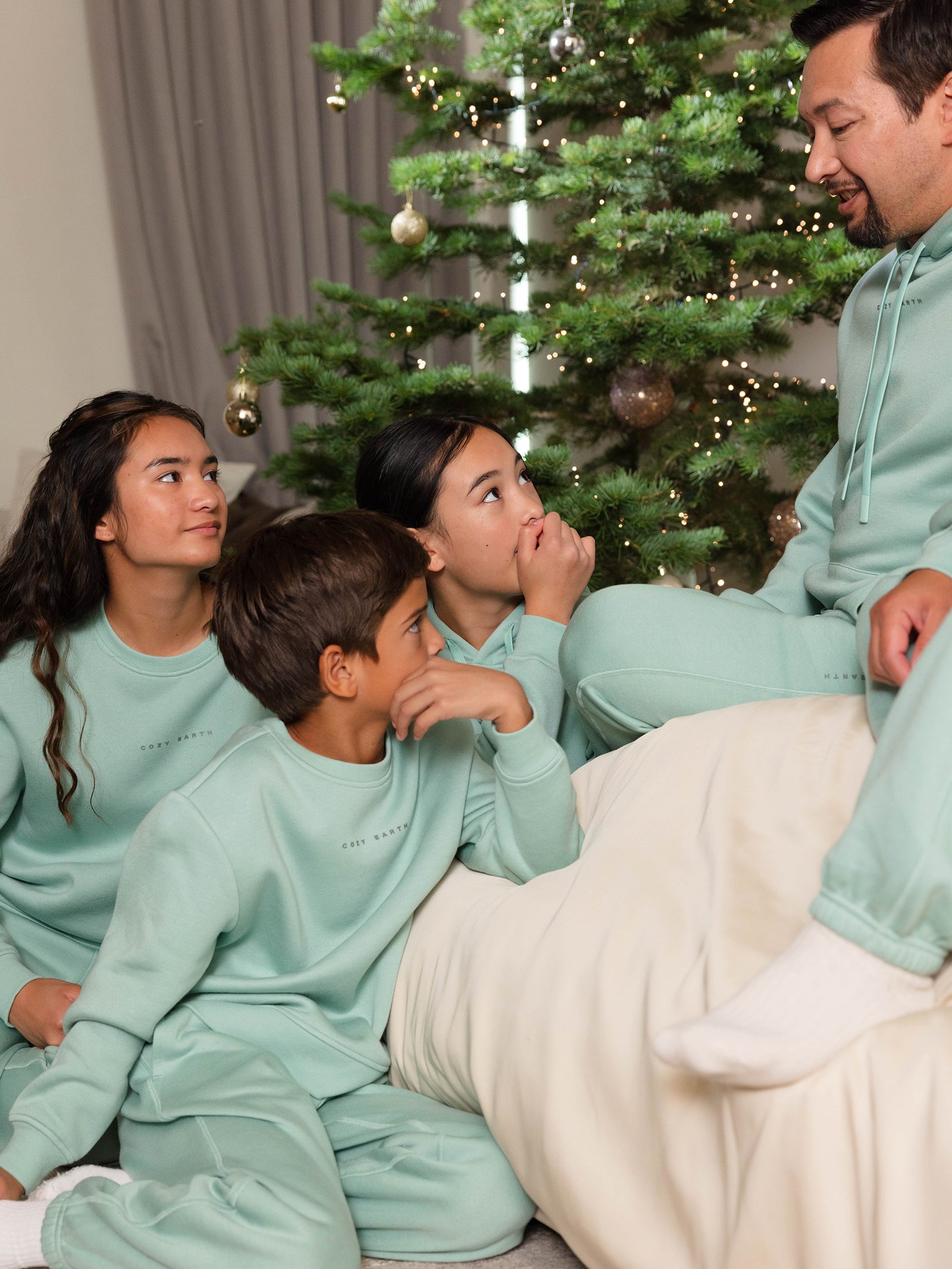 A family sitting together in matching Cozy Earth Kid's CityScape Crewnecks beside a decorated Christmas tree. The children are looking attentively at a man who is speaking, creating a cozy and festive atmosphere. 