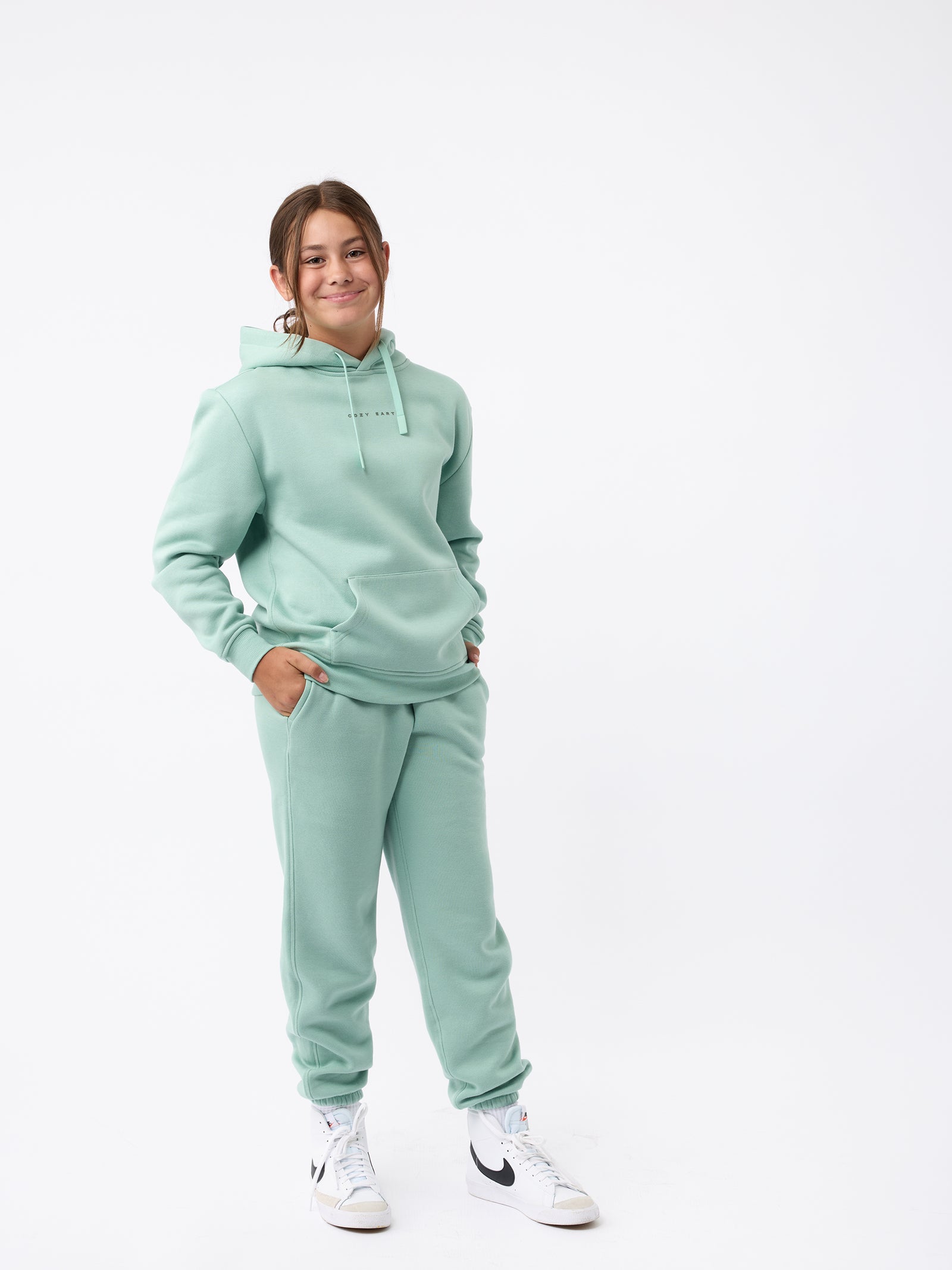 A person is wearing a Cozy Earth Kid's CityScape Hoodie in mint green along with matching sweatpants, standing with their hands in their pockets. They are smiling and have on white sneakers against a plain white studio background. 