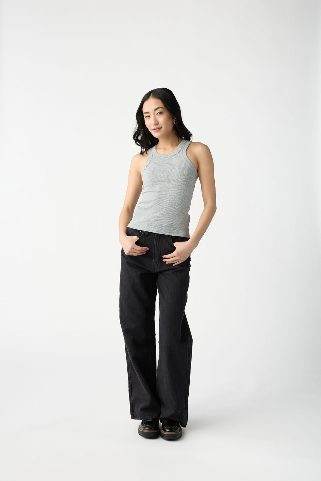 A dark haired woman wears Cozy Earth Women's Fine Ribbed Tank in Heather Grey. She hold the belt loops of her black jeans and stands in front of a white background. 