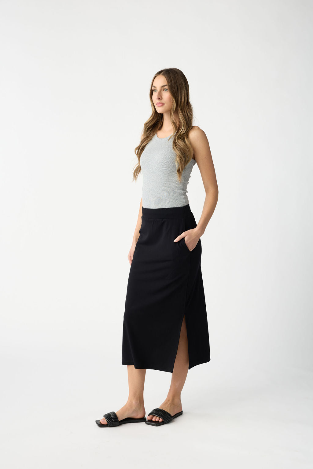 A woman poses against a plain white background, wearing Cozy Earth's Women's Brushed Bamboo Midi Skirt in black with side slits, paired with a gray sleeveless top and black sandals. She has long wavy hair and her hands in the skirt pockets. 