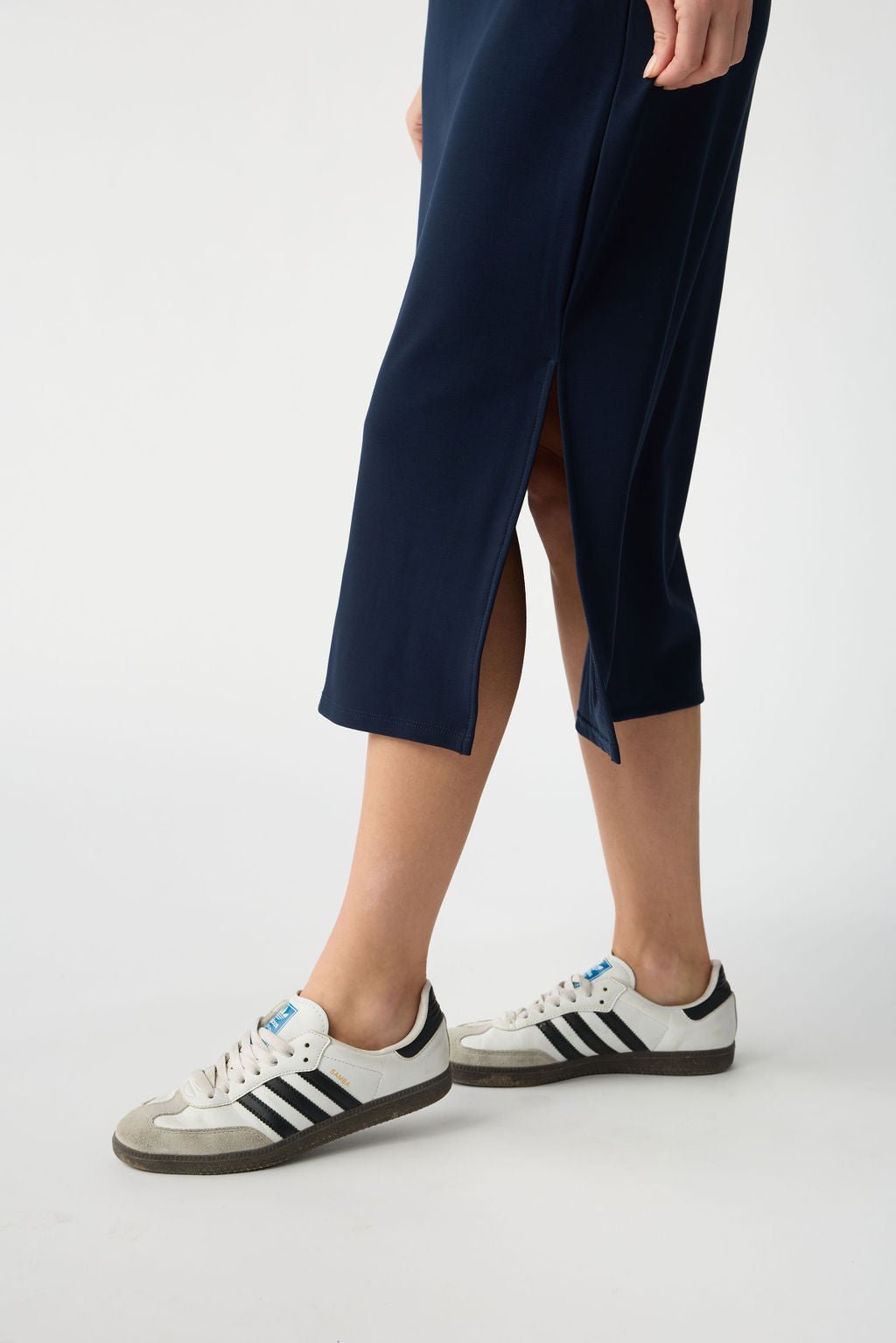 A person is wearing a dark blue skirt with a side slit and white Adidas sneakers featuring black stripes. The setting is plain and light-colored. 