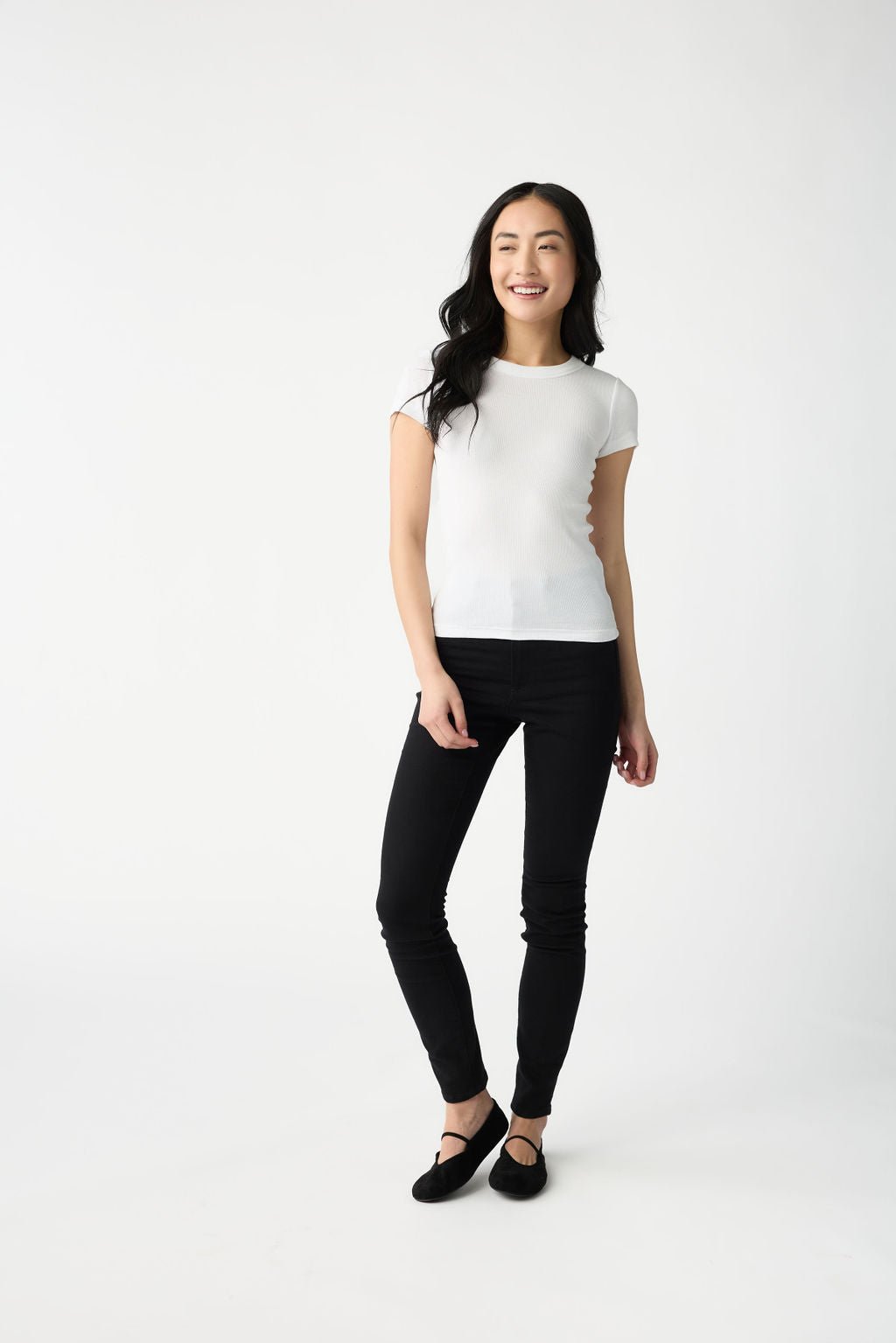 A dark haired woman wears Cozy Earth Women's Fine Ribbed Tee in White. She is wearing black pants and stands in front of a white background. 