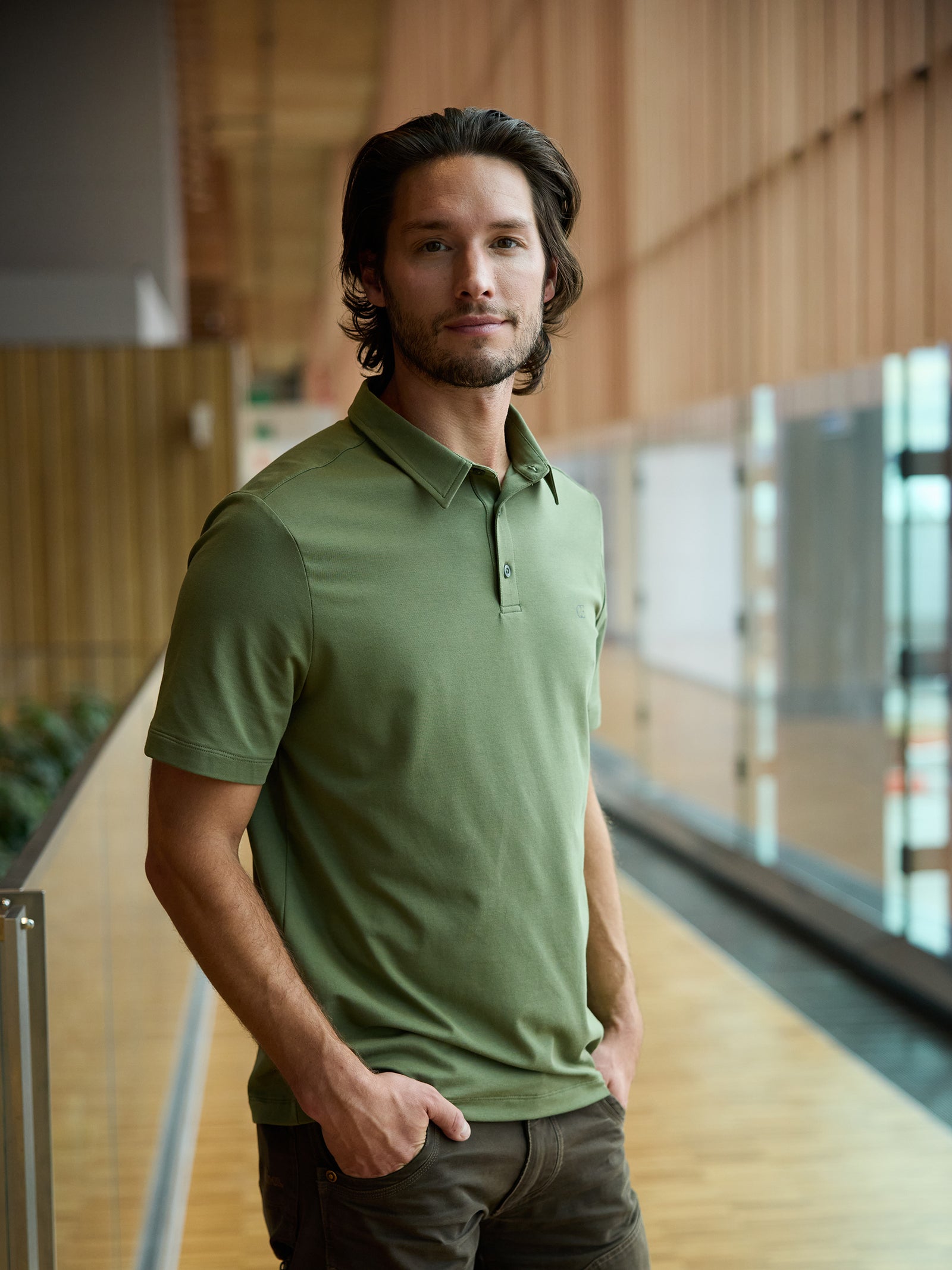 A person with shoulder-length hair stands in a corridor, dressed in a green Men's Everyday Polo by Cozy Earth and dark pants. The corridor features wooden paneling and glass walls. Smiling softly, the person has their hands in their pockets. 