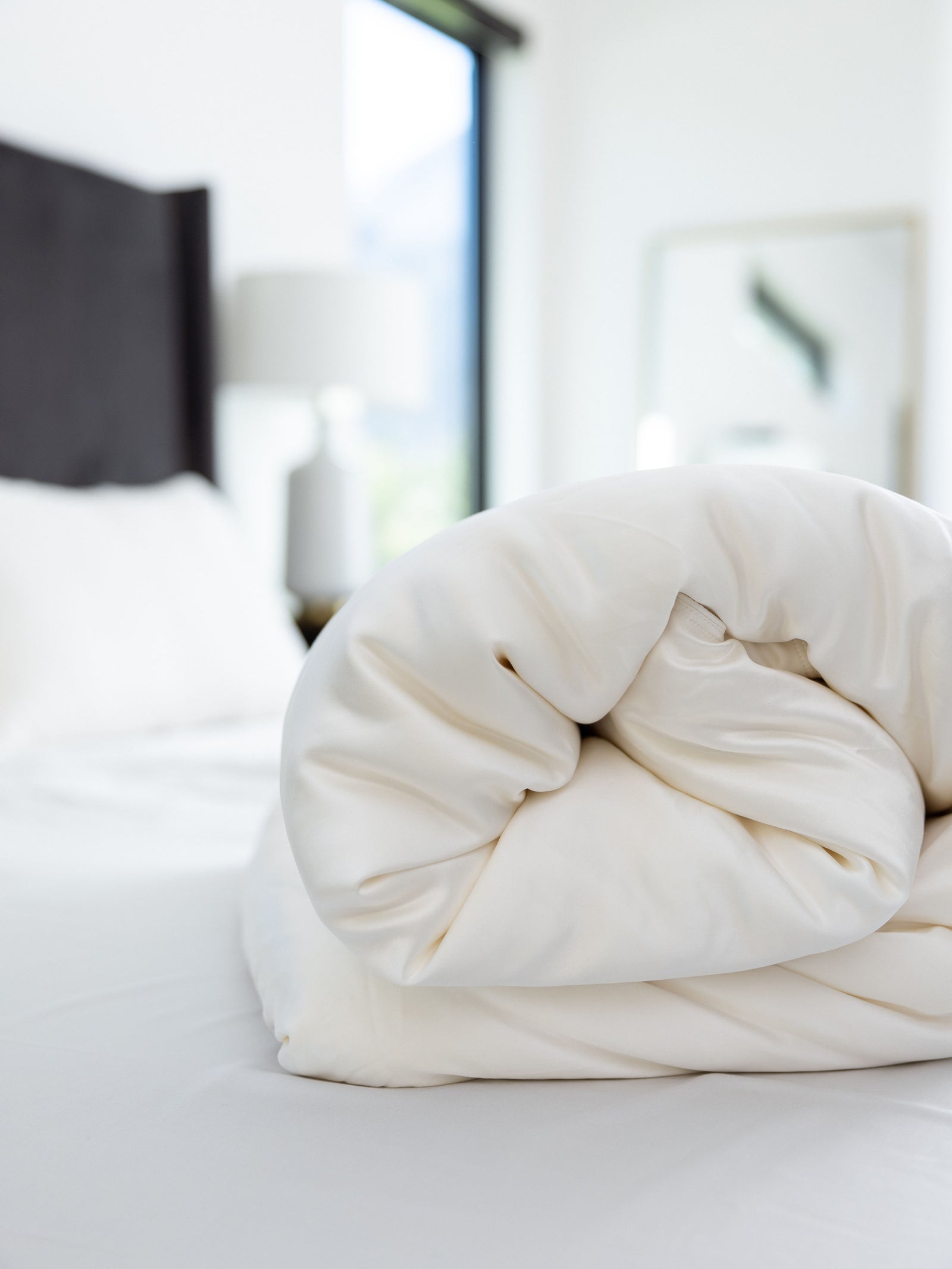 A neatly rolled-up Silk Comforter by Cozy Earth lies on a pristine white bed in a bright, modern bedroom. A black headboard, a lamp, and a framed picture in the background complement the room's minimalist decor.