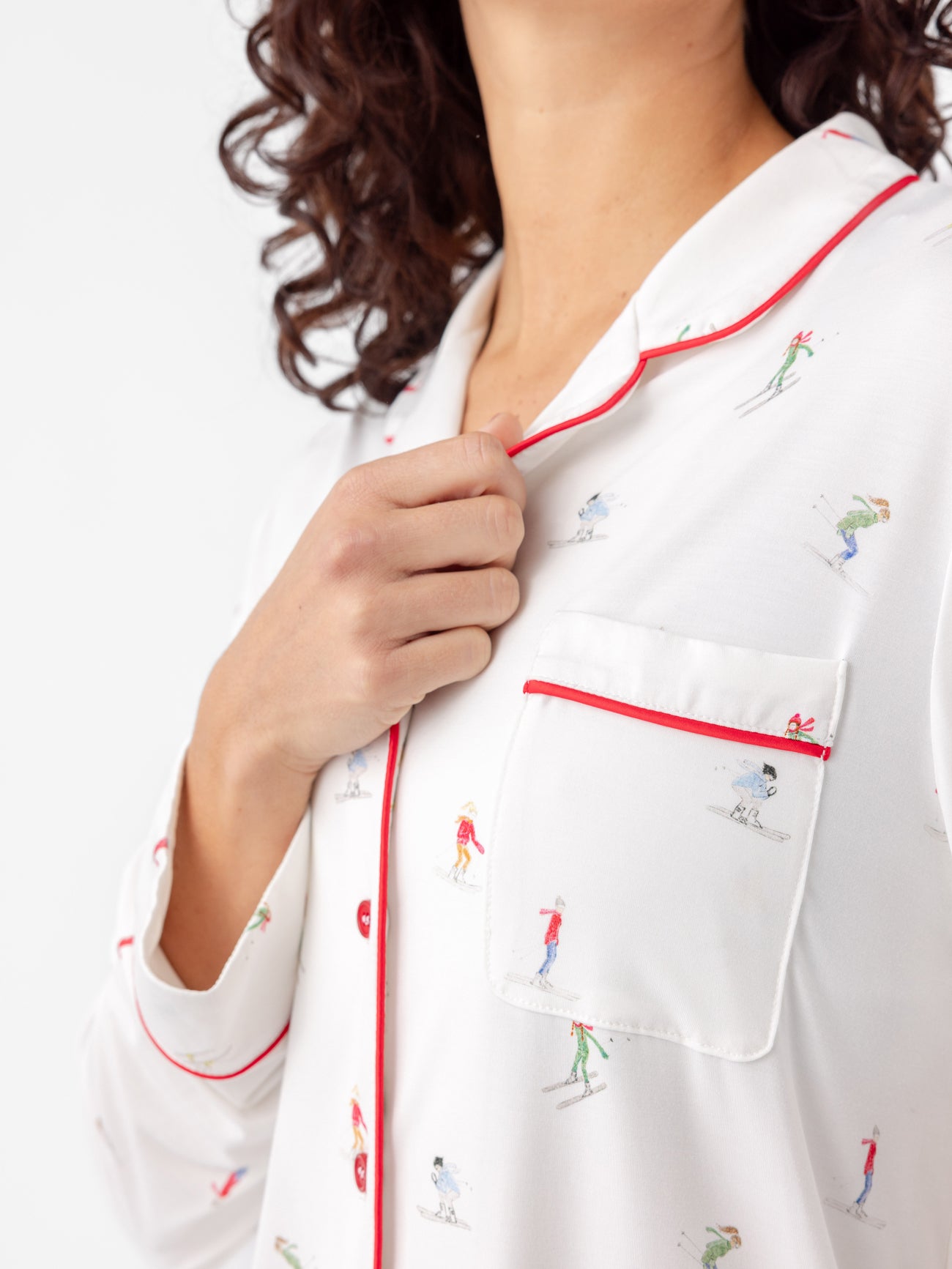 A person with curly hair models the Cozy Earth Last Chance Women's Bamboo Stretch-Knit Long Sleeve Pajama Set, showcasing a white top adorned with colorful skiing figures and featuring red piping on the collar and pocket. They are slightly pulling at the collar with their right hand. |Color:Skiers Ivory