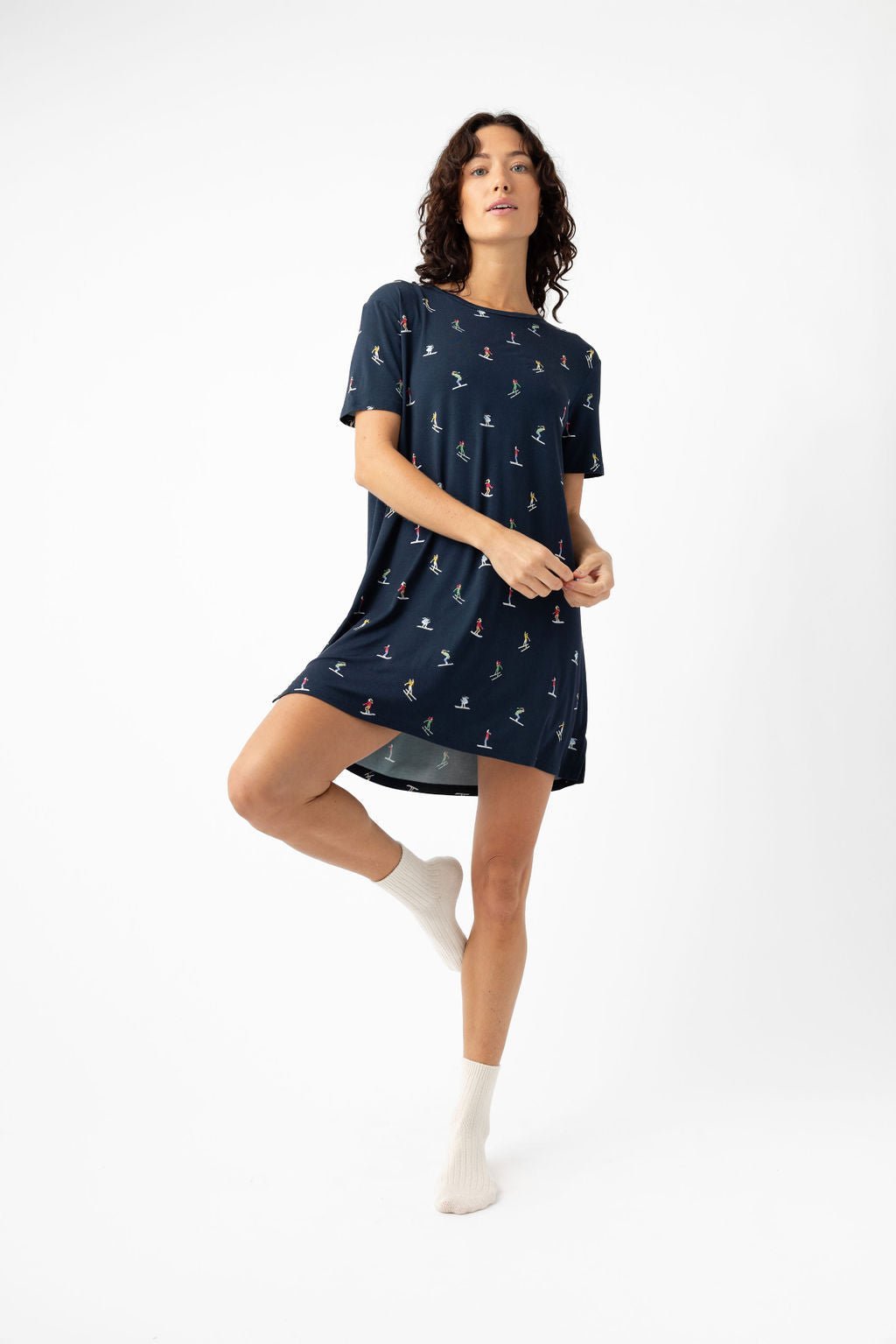 A woman stands on one leg against a white background, wearing the Cozy Earth Women's Bamboo Stretch-Knit Sleep Dress with small colorful prints and white socks. She looks slightly upward, holding the edge of her dress with one hand. 