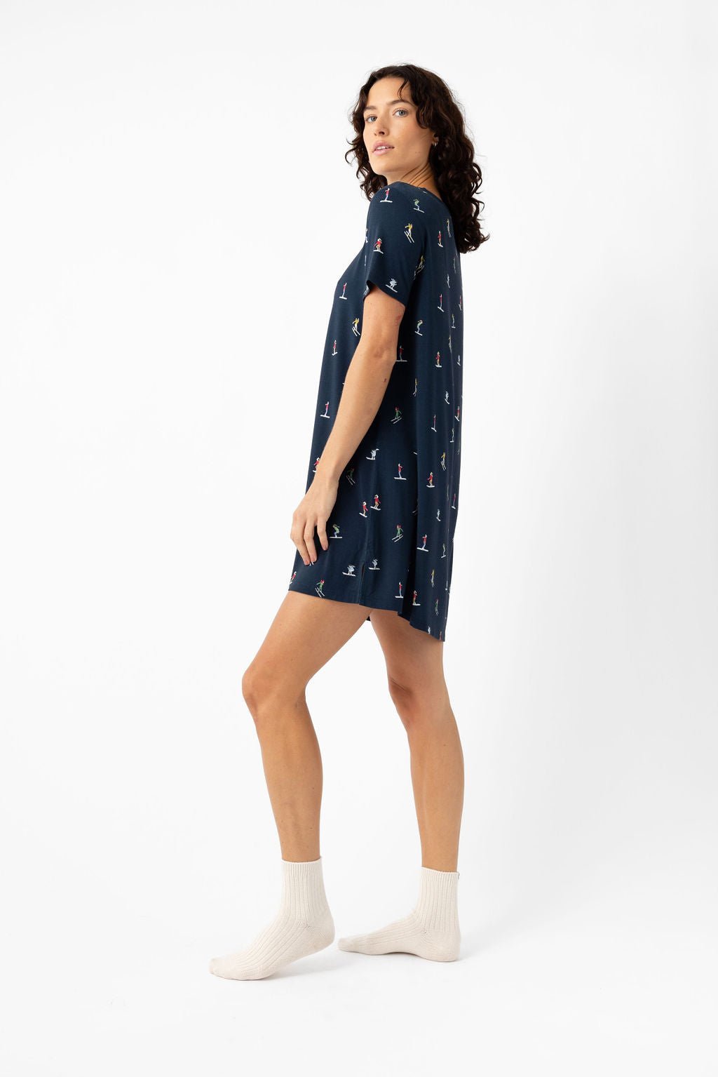A woman with curly hair stands sideways in a Cozy Earth Women's Bamboo Stretch-Knit Sleep Dress, featuring a navy blue color with small sailboat prints and white ankle socks, against a plain white background. 