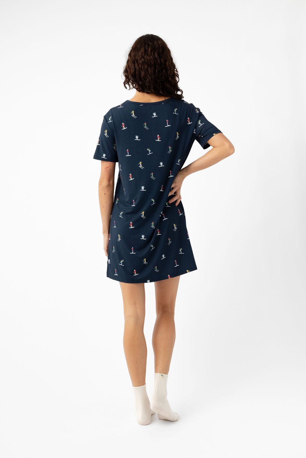 A woman with curly hair stands with her back to the camera, wearing the Cozy Earth Women's Bamboo Stretch-Knit Sleep Dress in navy blue with a small colorful print and white ankle socks, against a plain white background. 