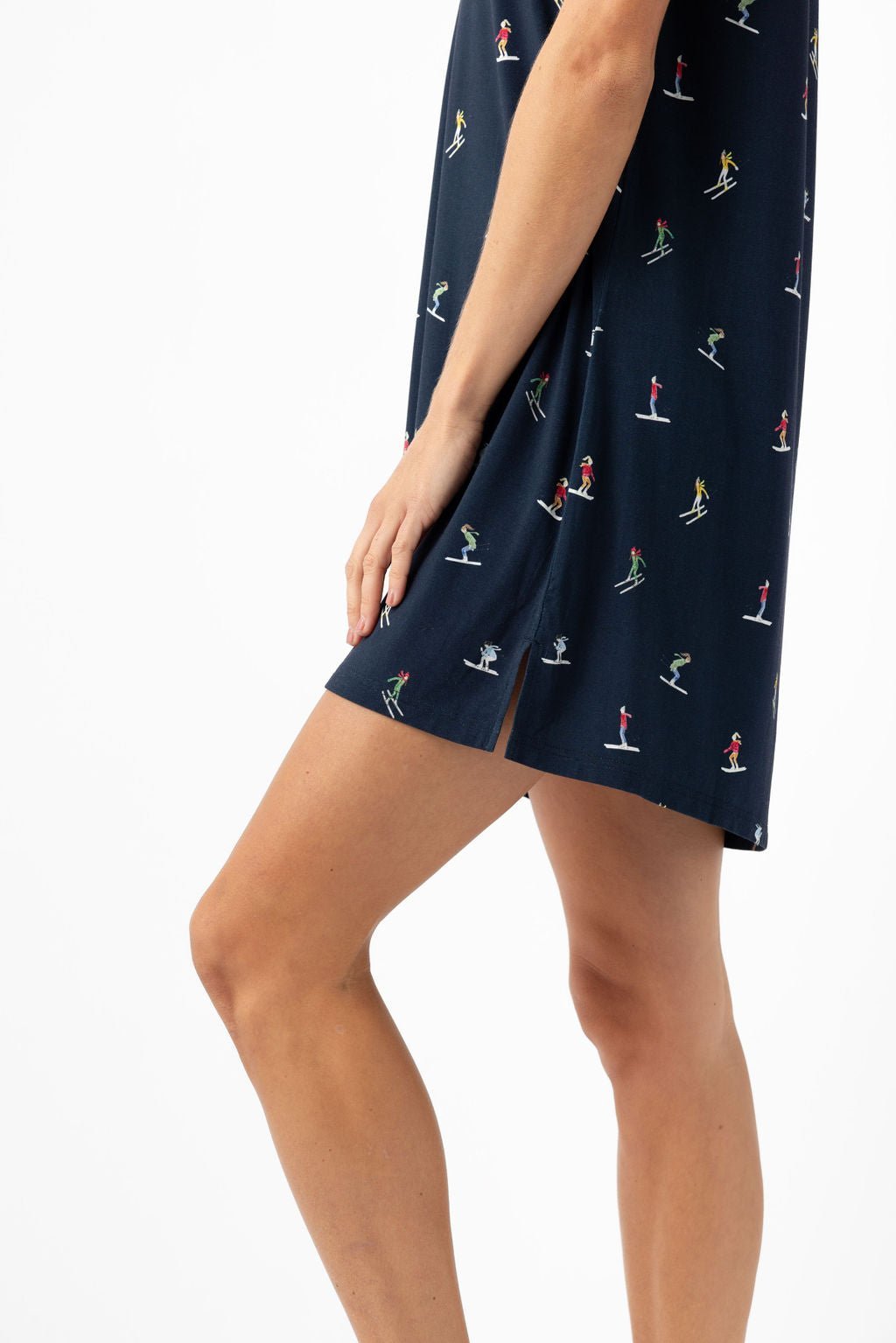 A person wearing the Cozy Earth Women's Bamboo Stretch-Knit Sleep Dress in navy blue with small, colorful skier prints stands against a white background. The dress has a small slit near the hem and only the lower half of their body is visible. 