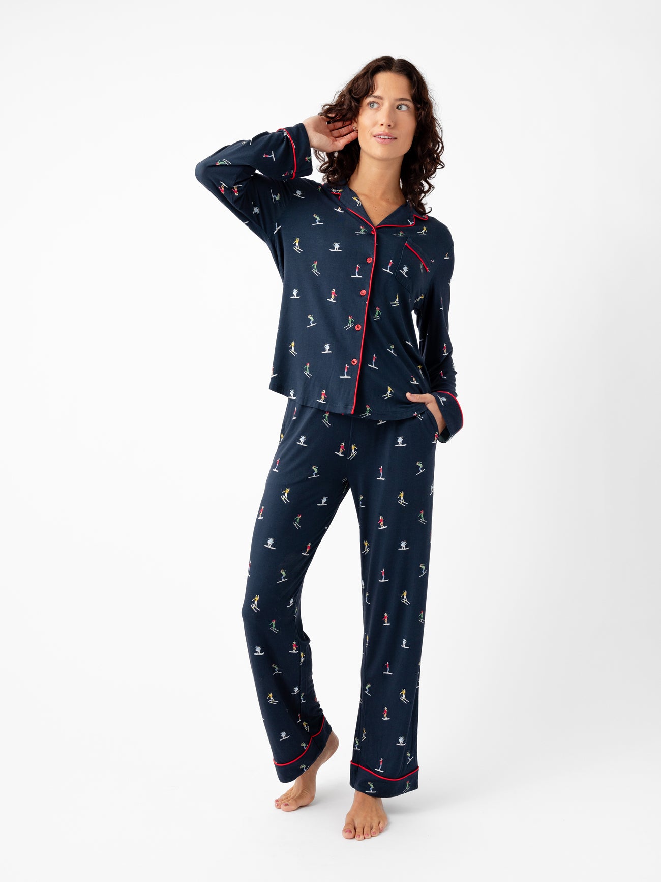 A person stands barefoot in Cozy Earth's Last Chance Women's Bamboo Stretch-Knit Long Sleeve Pajama Set, featuring a navy design with colorful animal prints. The collared top has buttons and red piping on the cuffs and edges, paired with full-length pants against a plain white background. |Color:Skiers Navy