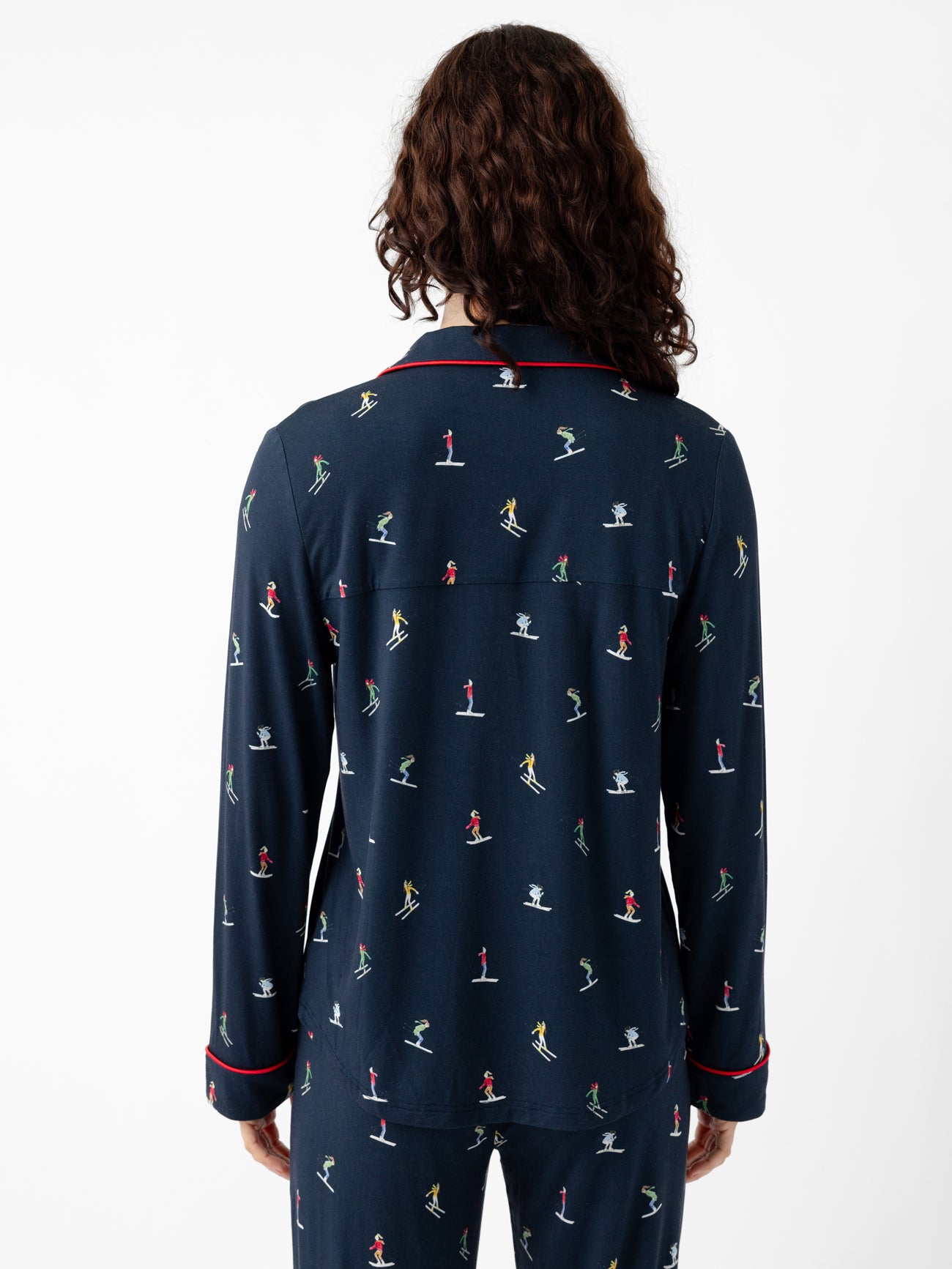 A person with long, curly hair stands facing away, wearing Cozy Earth's Last Chance Women's Bamboo Stretch-Knit Long Sleeve Pajama Set featuring small colorful figures and red piping on the collar and cuffs. The set comes with matching bottoms, all set against a plain white background. 