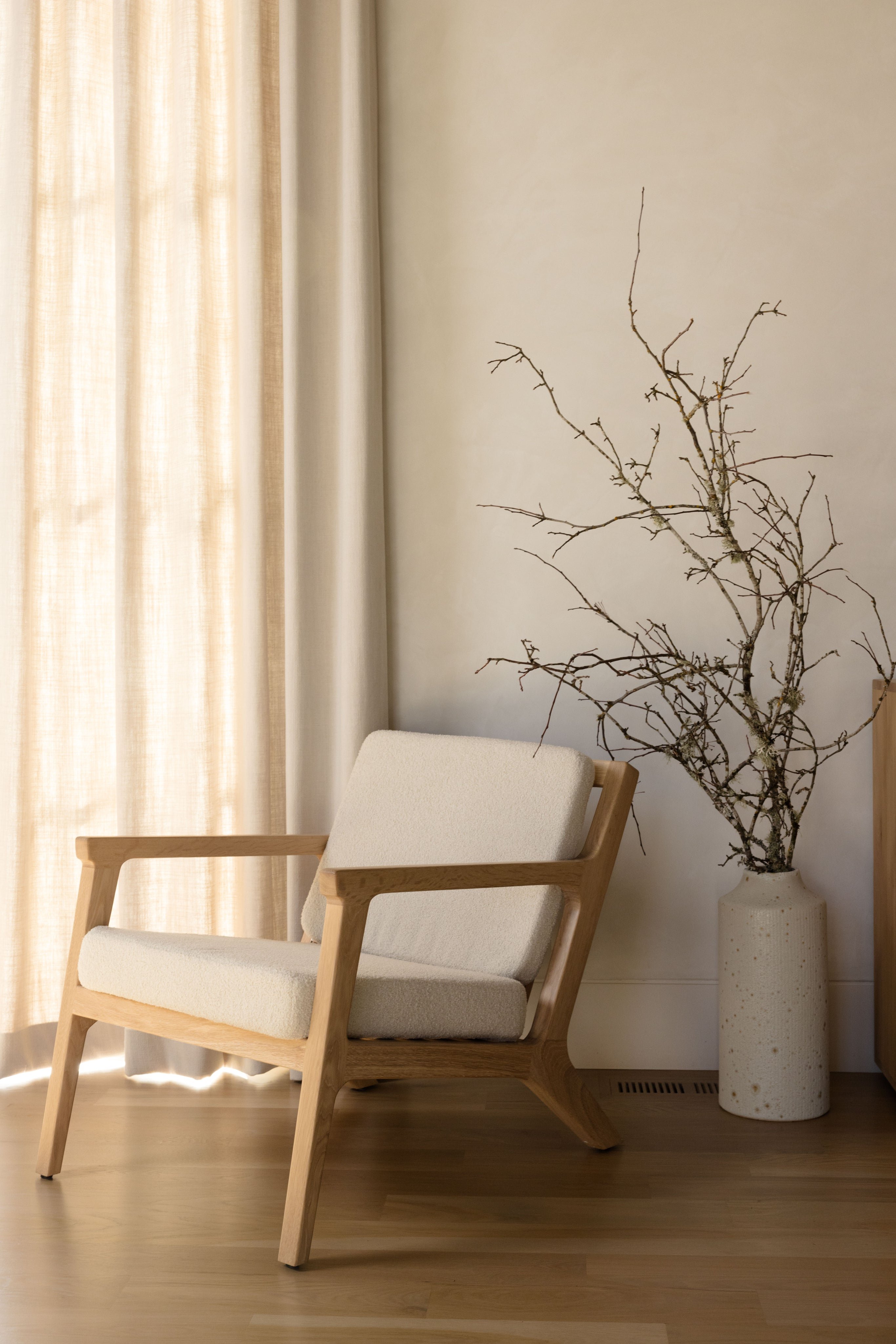 A minimalist living space featuring a light wooden armchair with cream cushions. Beside it, a vase holds bare branches. Soft sunlight filters through sheer curtains, casting gentle shadows on the floor.