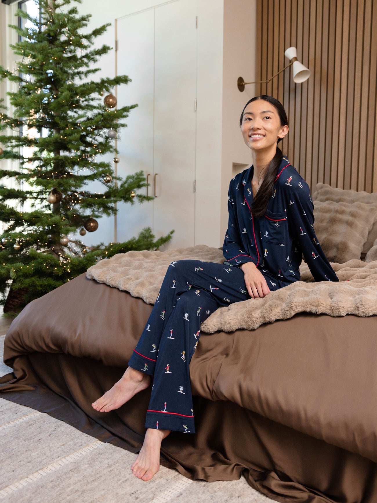 A person wearing Cozy Earth's Last Chance Women's Bamboo Stretch-Knit Long Sleeve Pajama Set in dark blue sits smiling on the bed's edge. The cozy room features a fluffy blanket and a decorated Christmas tree in the background. 