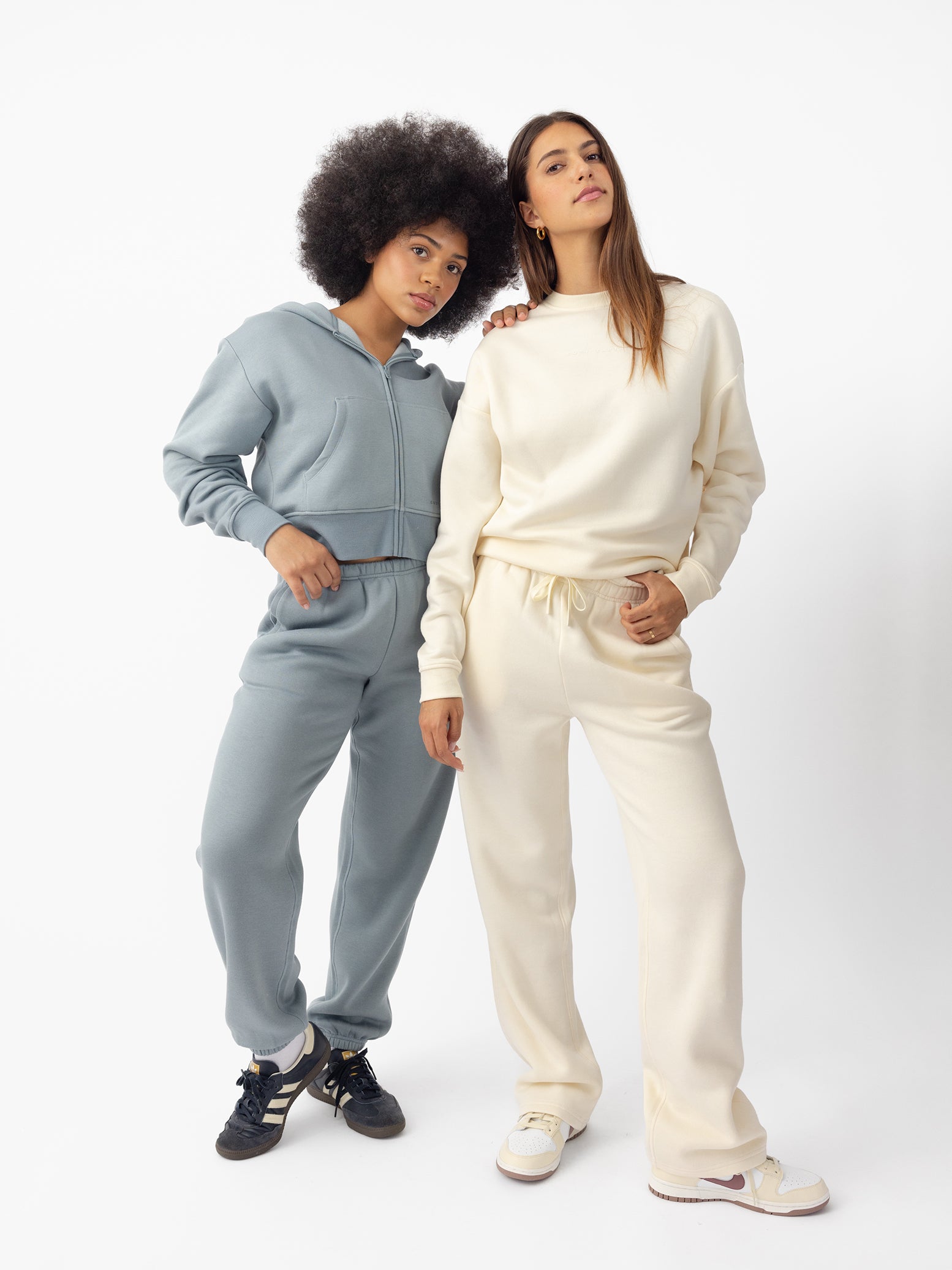 Two women wear Cozy Earth CityScape apparel; one in the Cropped Full-Zip Hoodie and Sweatpants, the other in the Crewneck Sweatshirt and Wide Leg Pants.