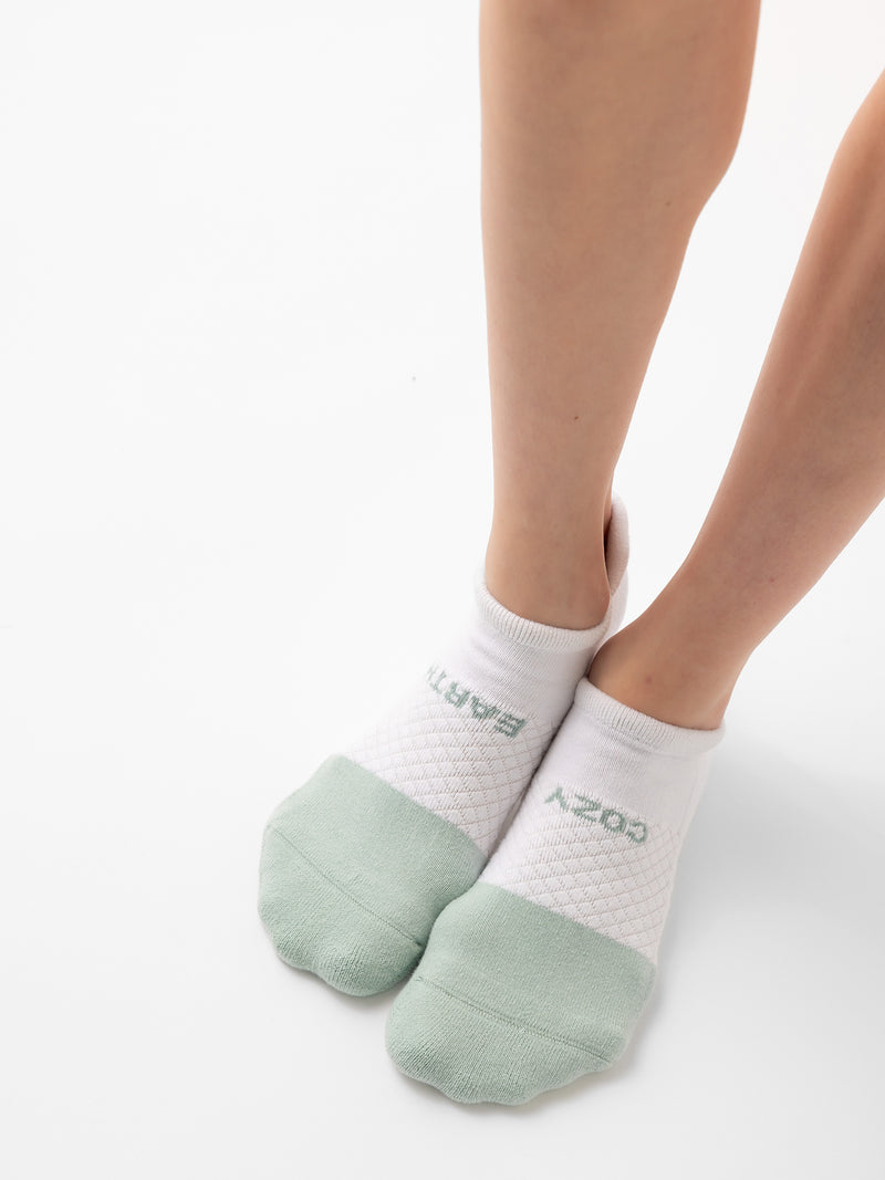 A person wearing Cozy Earth's Essential Ankle Socks from the 4-pack, featuring white material with green toe caps and the words "BAST" and "CORE" on them. The image focuses on their legs and feet against a plain white background.