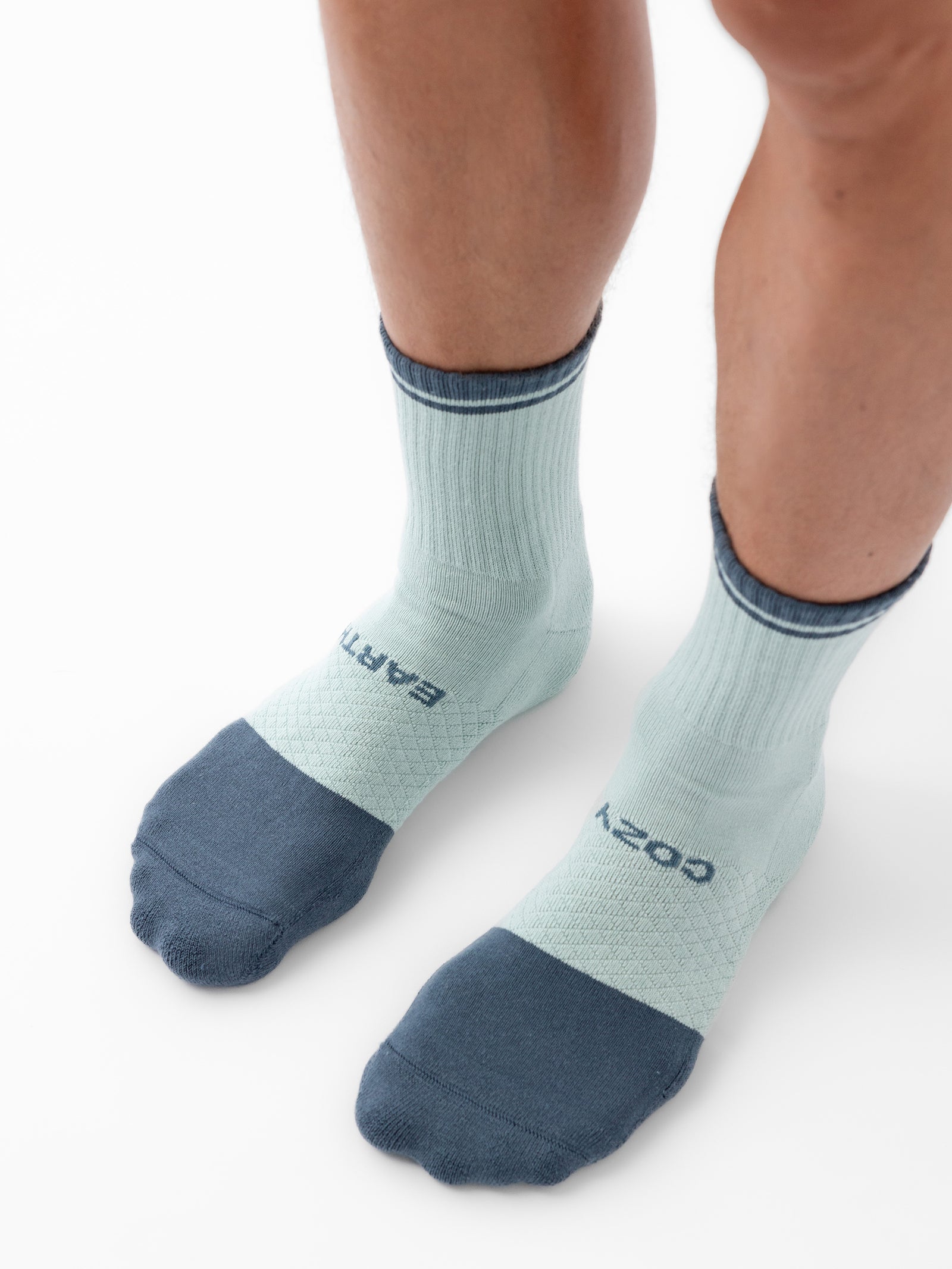 Close-up of a pair of legs wearing Cozy Earth's Essential Calf Socks in light green with dark blue reinforced toes and an elastic band at the top. The socks feature text near the ankle area. The white background emphasizes the socks. 