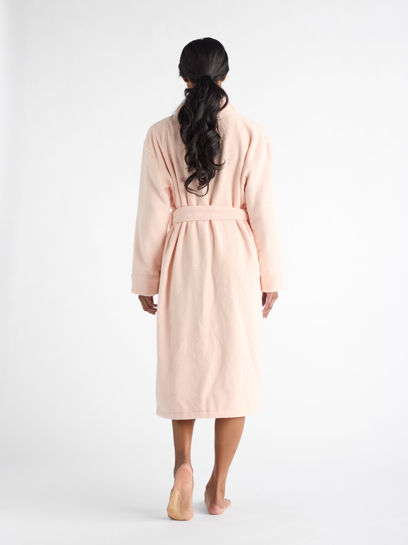 A person is standing barefoot, facing away, wearing a Luxe Bath Robe by Cozy Earth in light pink. The robe is belted at the waist, and the person's long, dark hair hangs down their back against a plain white background. 