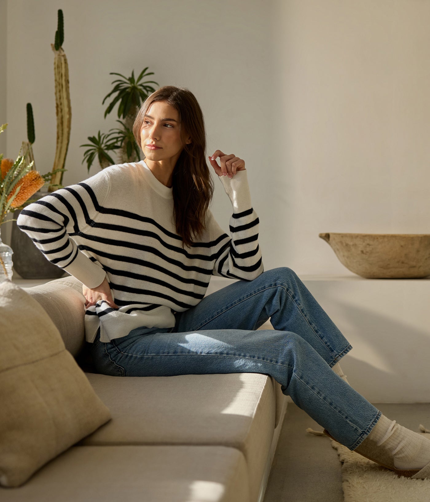 A woman wearing a Cozy Earth Women's Rowan Sweater and jeans sits on a beige couch in a bright room with plants and a wooden bowl on the table, gazing thoughtfully at the window as natural light illuminates the scene. 