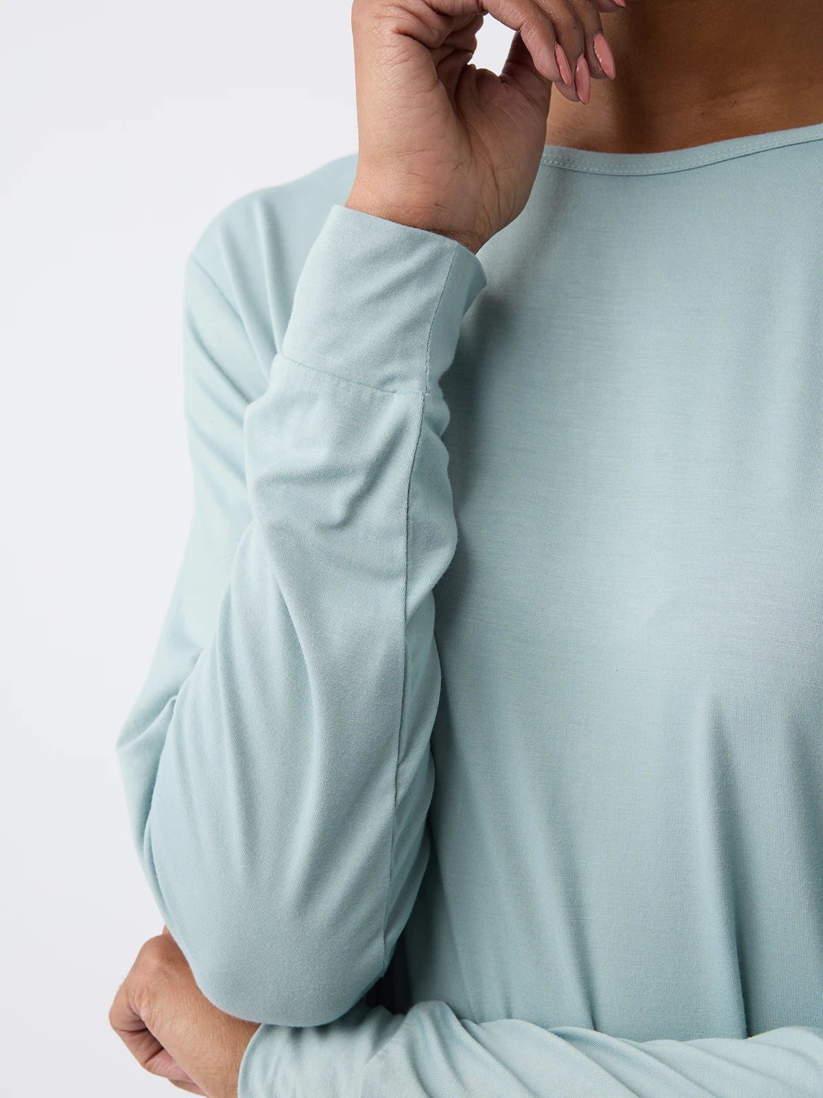 A woman wearing the Cozy Earth Women's Stretch Knit Bamboo Long Sleeve Lounge Tee in light blue gently touches their face with light pink painted nails. The shirt's fabric looks soft, set against a plain, neutral background. 